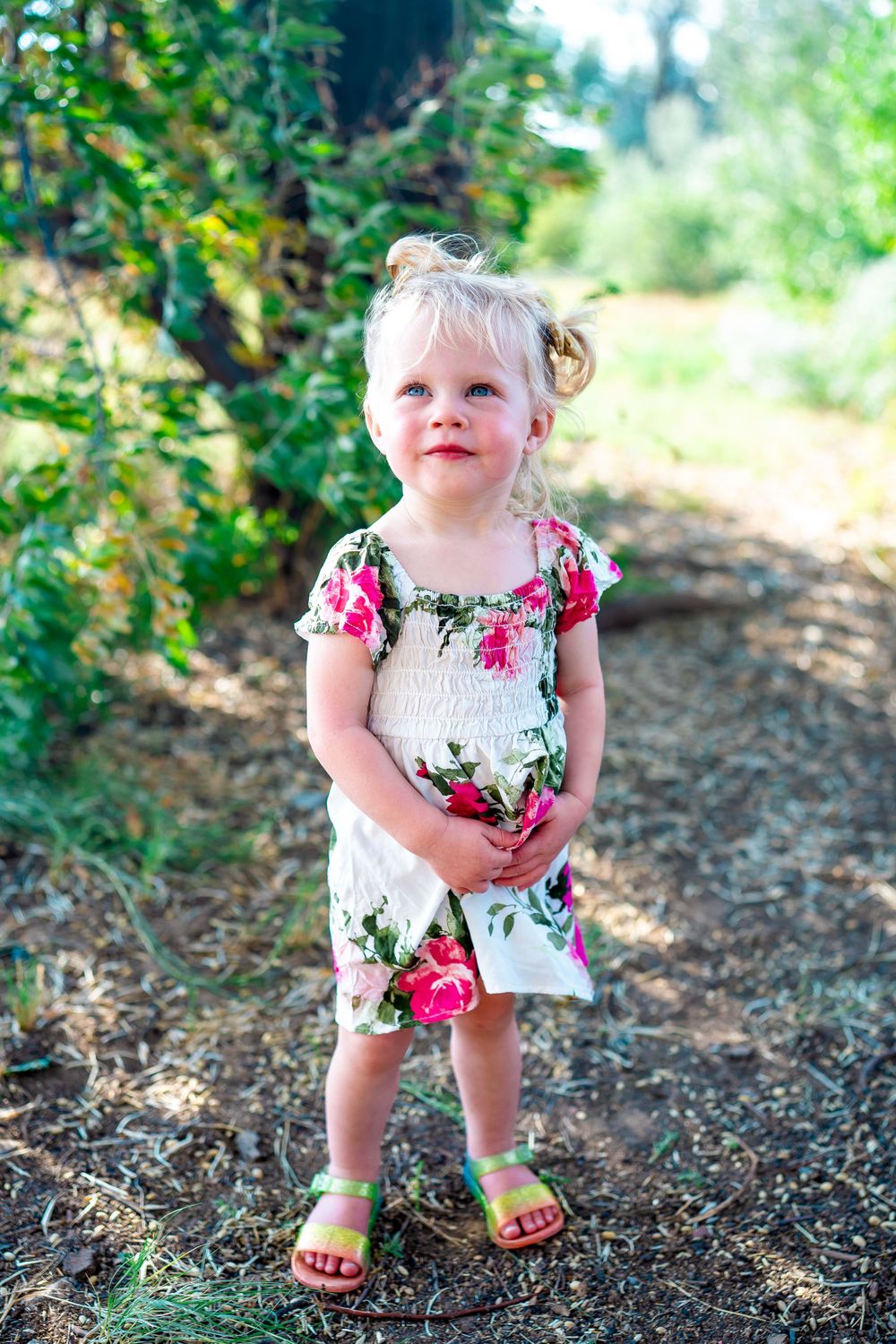 Toddler wearing floral dress and yellow sandals stands on outdoor trail.