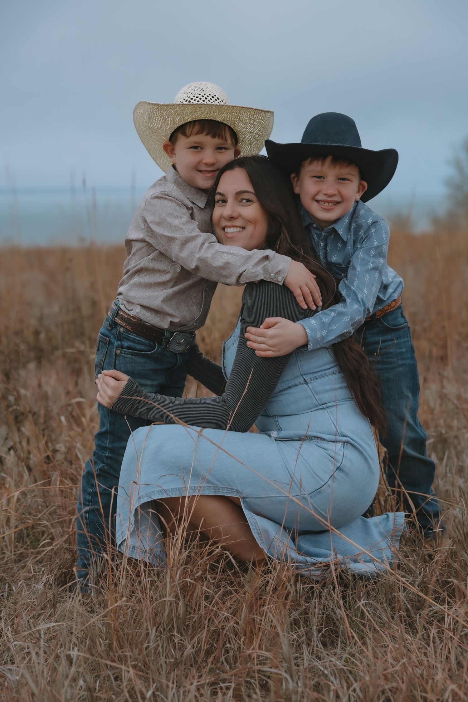 A smiling mother kneels in a field hugged by two young boys wearing cowboy hats and denim outfits in Garrison, ND before going to Bismarck