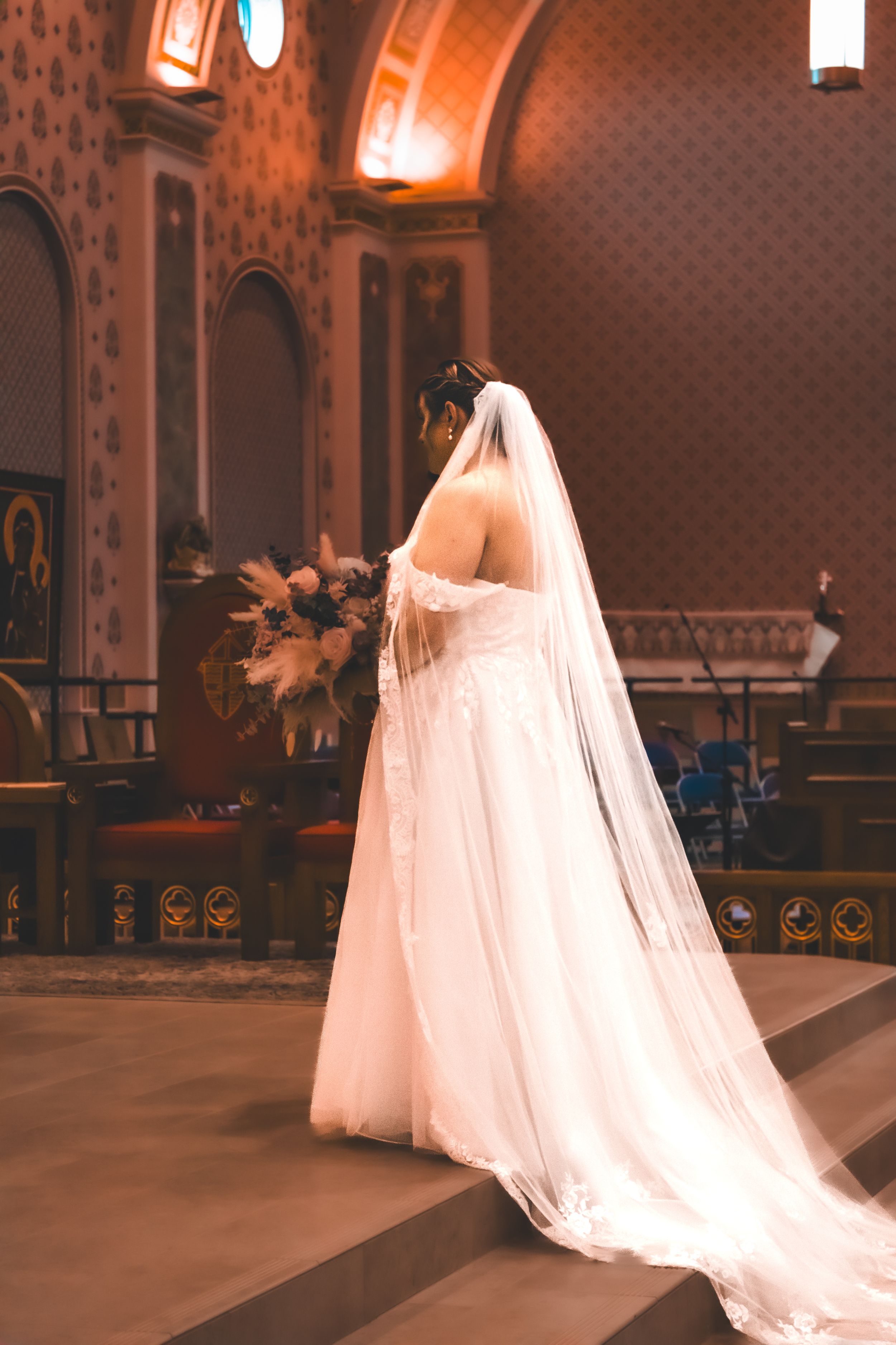 Bride standing alone at the church altar, holding her bouquet, framed by the soft light filtering through the space.