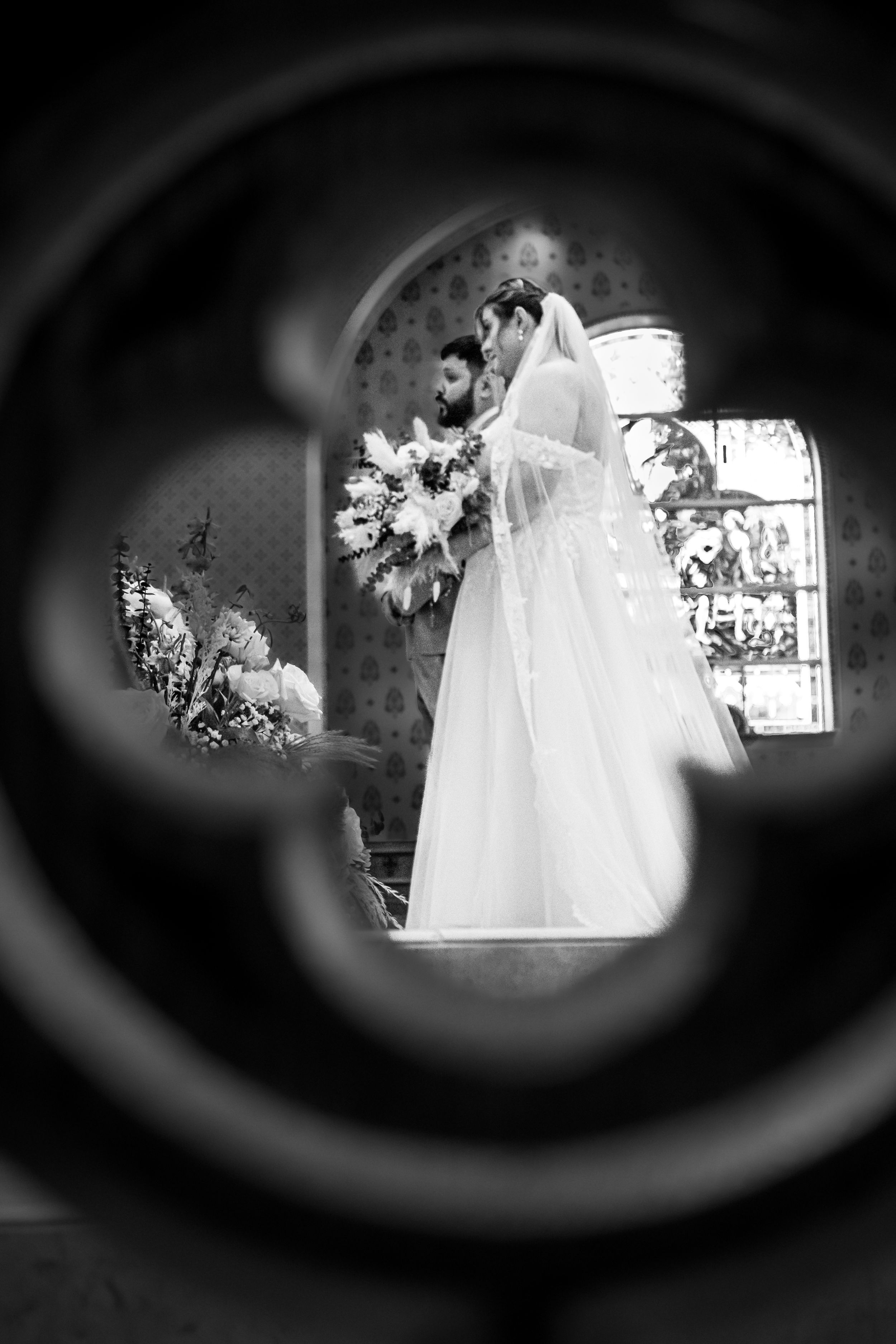 A bride and groom standing at the altar, viewed through the blurred outline of a church door keyhole, creating a romantic, intimate frame.