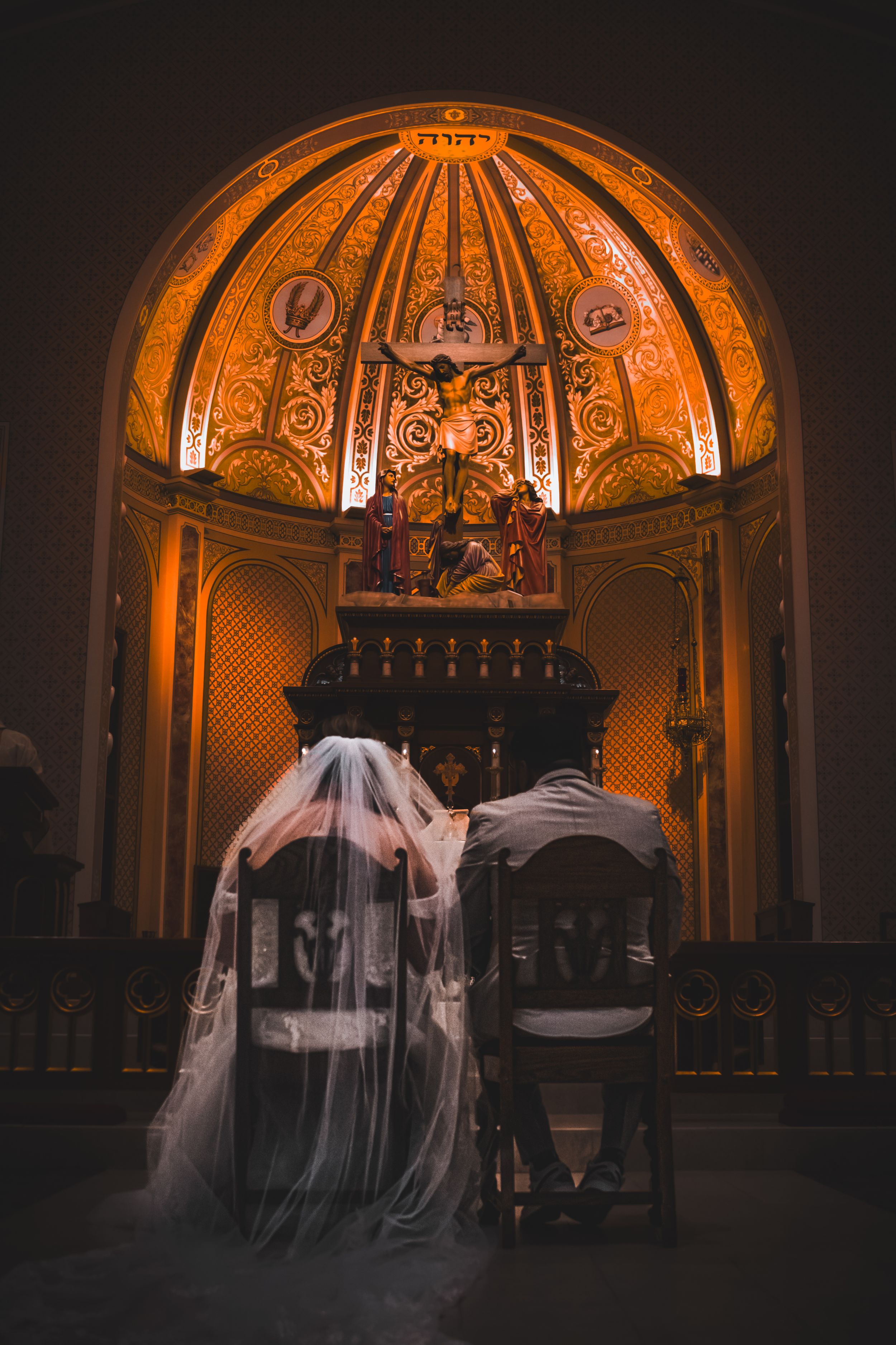 Bride and groom seated side by side in chairs, with their backs to the camera, facing the church altar in a quiet, reflective moment.