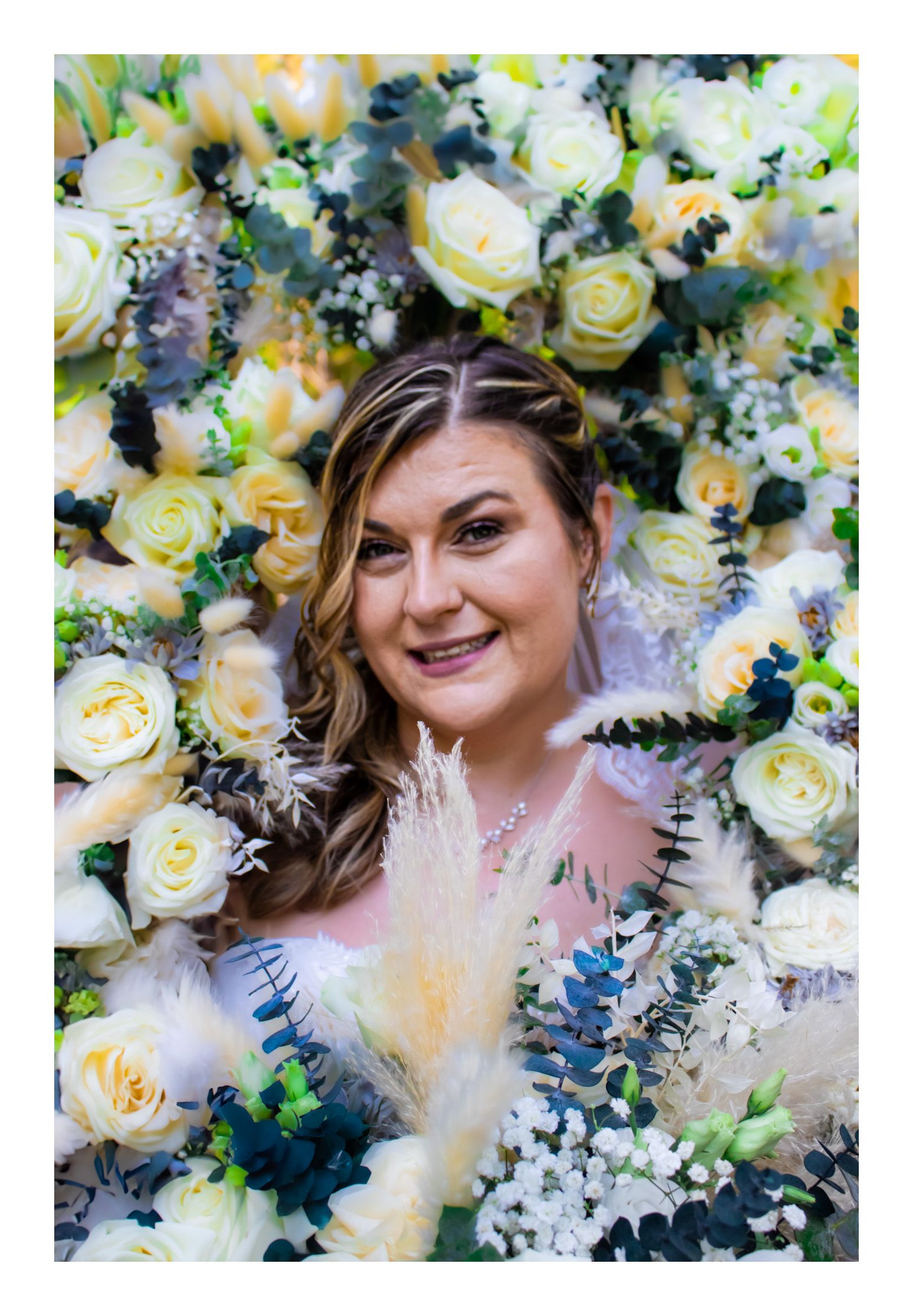 Bride smiling brightly, her head surrounded by bouquets held by her bridal party, with soft natural light illuminating the joyful scene.