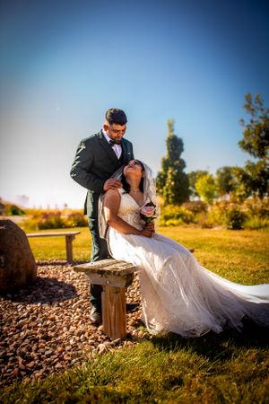 Wedding couple are posed outdoors in a grassy area.  The bride is sitting on a bench with the groom standing behind her.