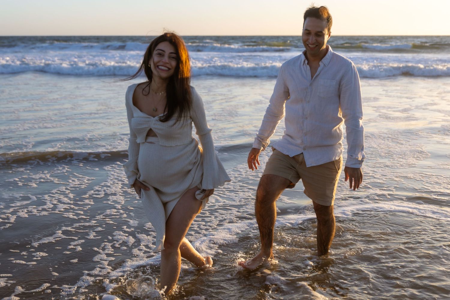 Color portrait of a smiling pregnant woman and her husband wading through shallow water in the ocean.