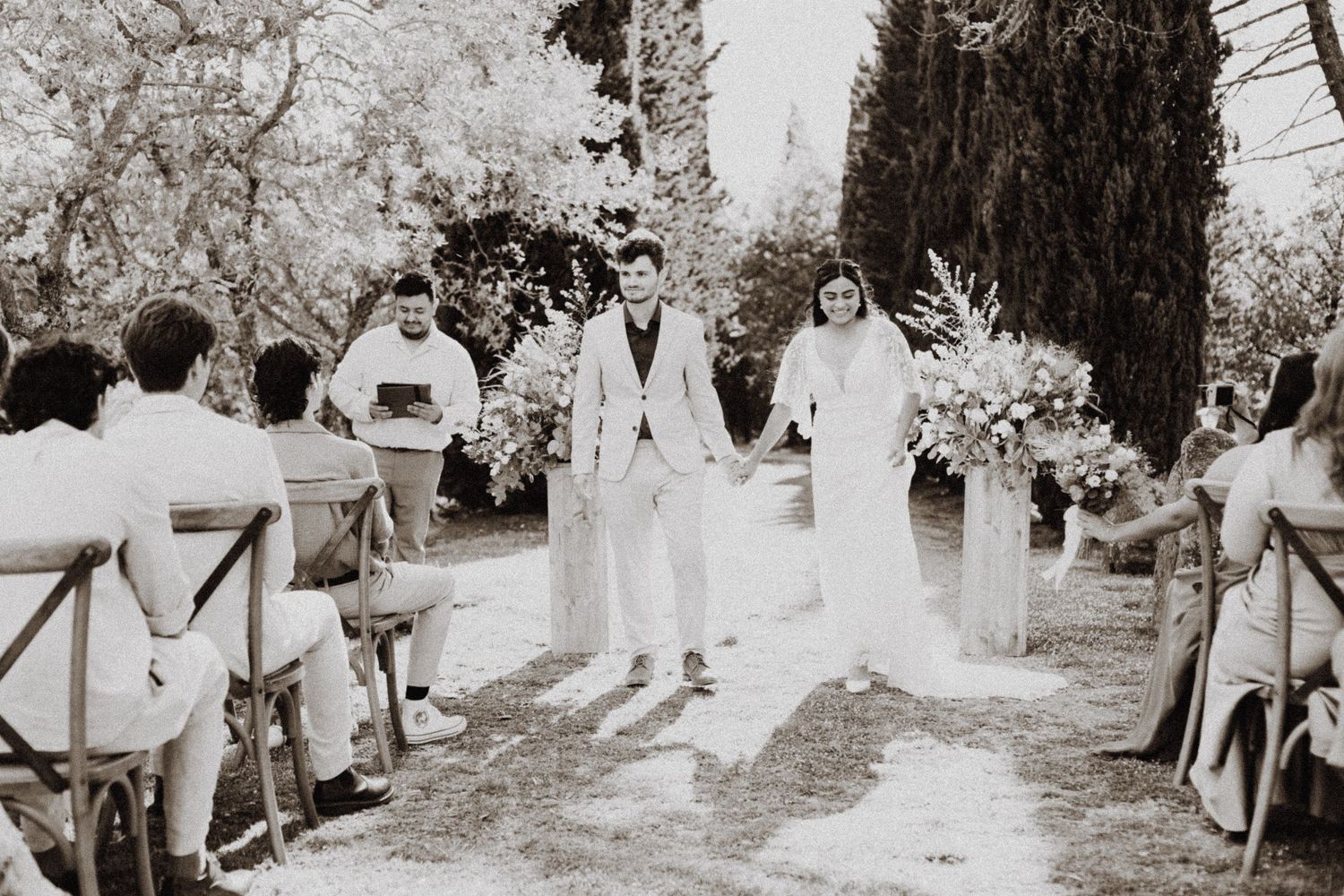 A black and white photograph of an outdoor wedding ceremony with cypress trees lining the aisle.