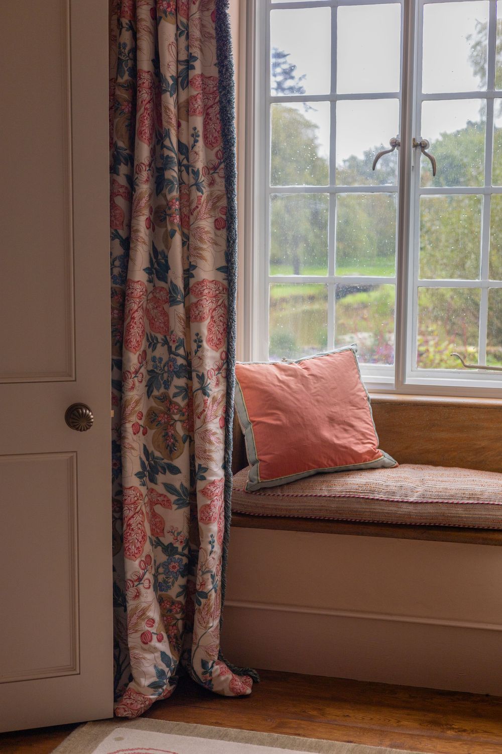 Cozy window seat with coral pillows and floral curtains overlooking a pastoral landscape.