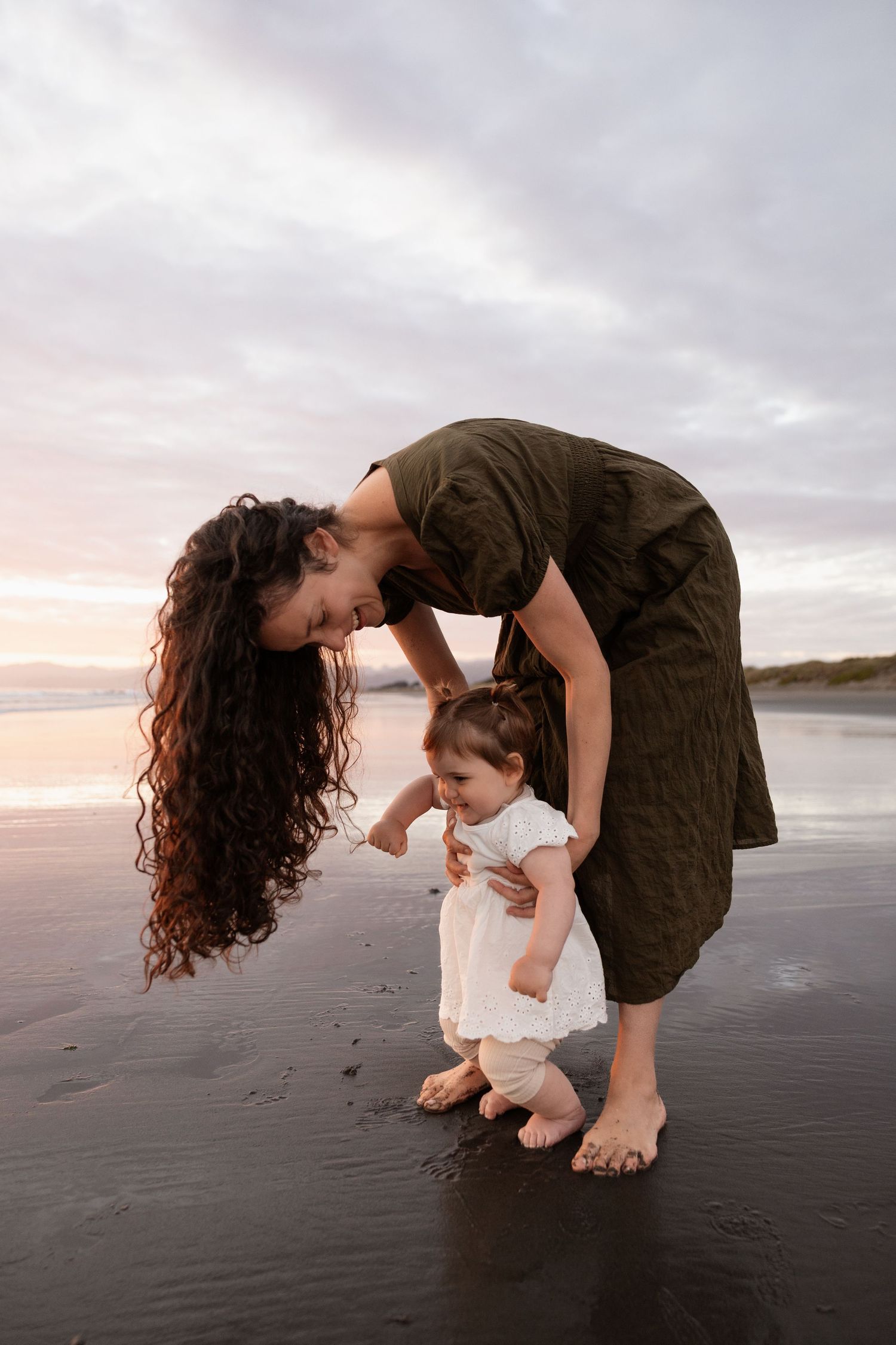 mum leaning over her baby and holding onto her at the beach during sunrise family session christchurch