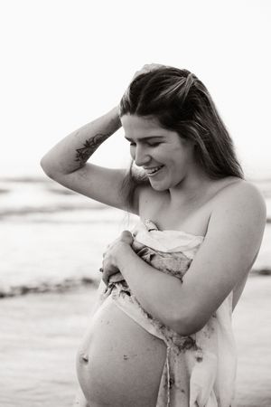mum to be holding onto a sheet wrapped around her, looking and smiling at baby bump during sunrise maternity session in christchurch