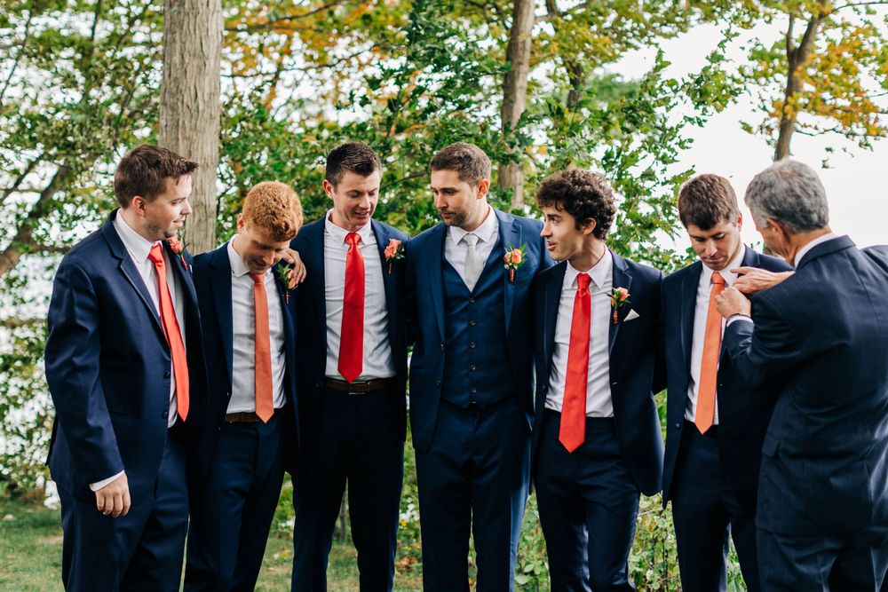Wedding party of groomsmen share a candid laughing moment in navy suits and red ties.