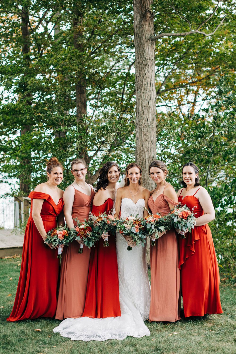 Wedding party portrait featuring bridesmaids in shades of red and coral with lush greenery backdrop.