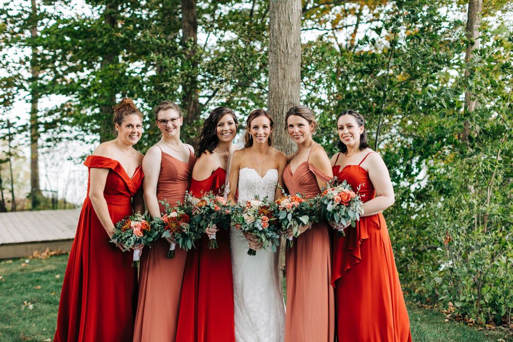 Bridesmaids in coral and red dresses hold bouquets while posing with the bride outdoors.
