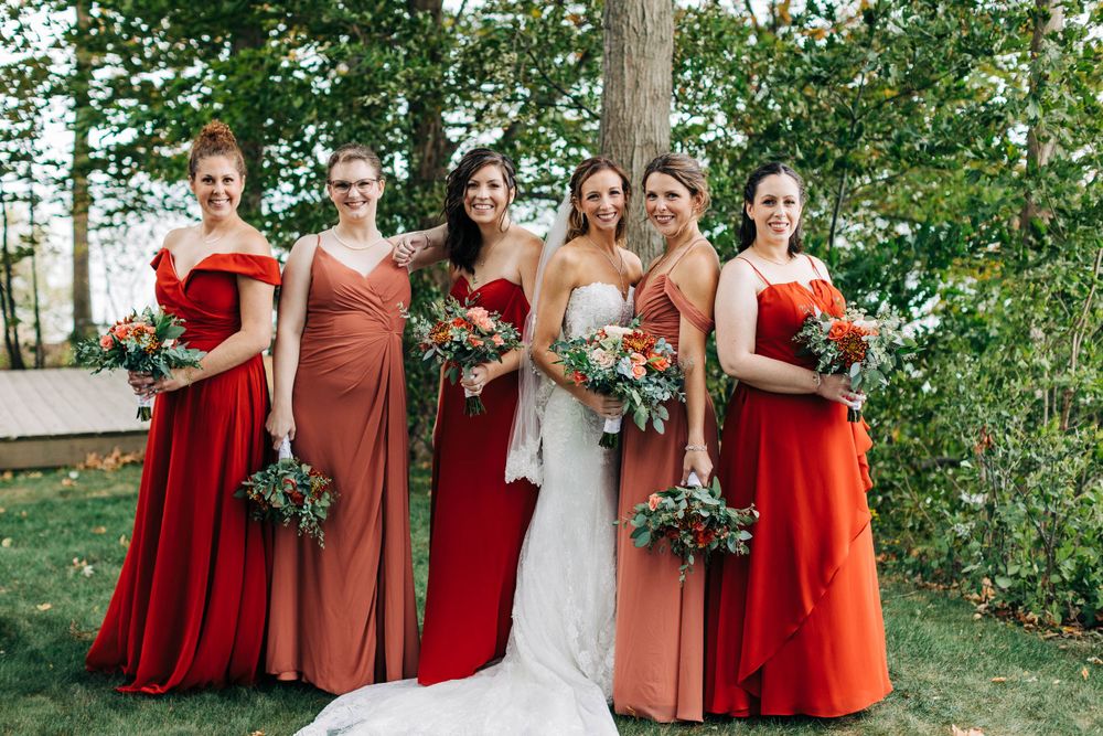 Wedding party poses outdoors wearing coordinating red and coral dresses with floral bouquets.