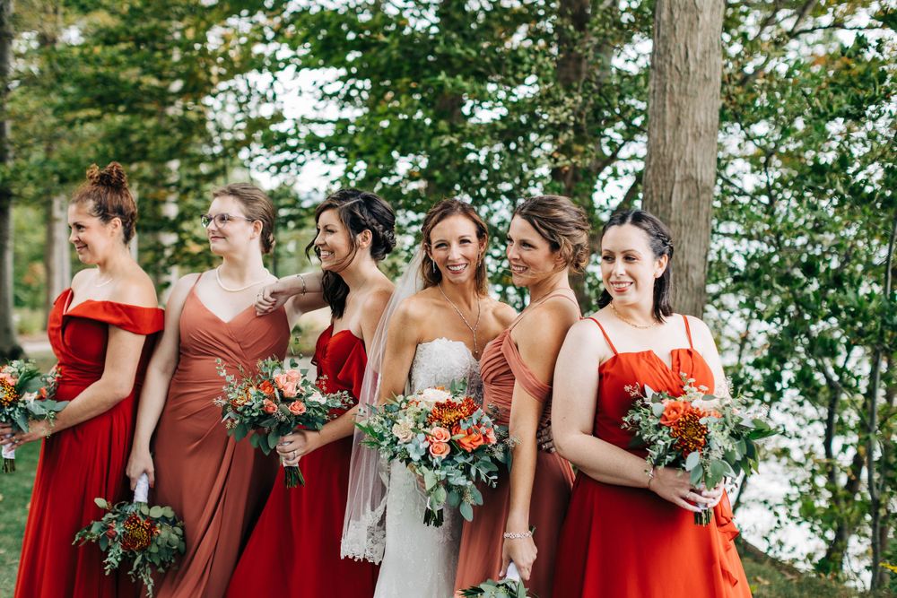 Wedding party in shades of red and coral dresses holding vibrant autumn bouquets against a green backdrop.