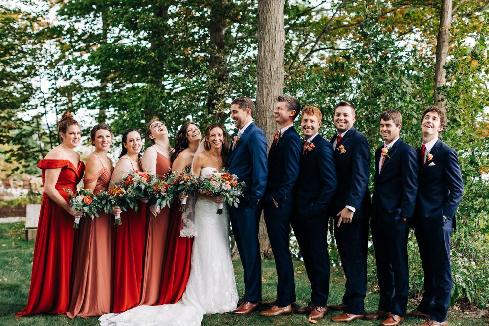 Wedding party in navy suits and red dresses poses together outdoors under trees.