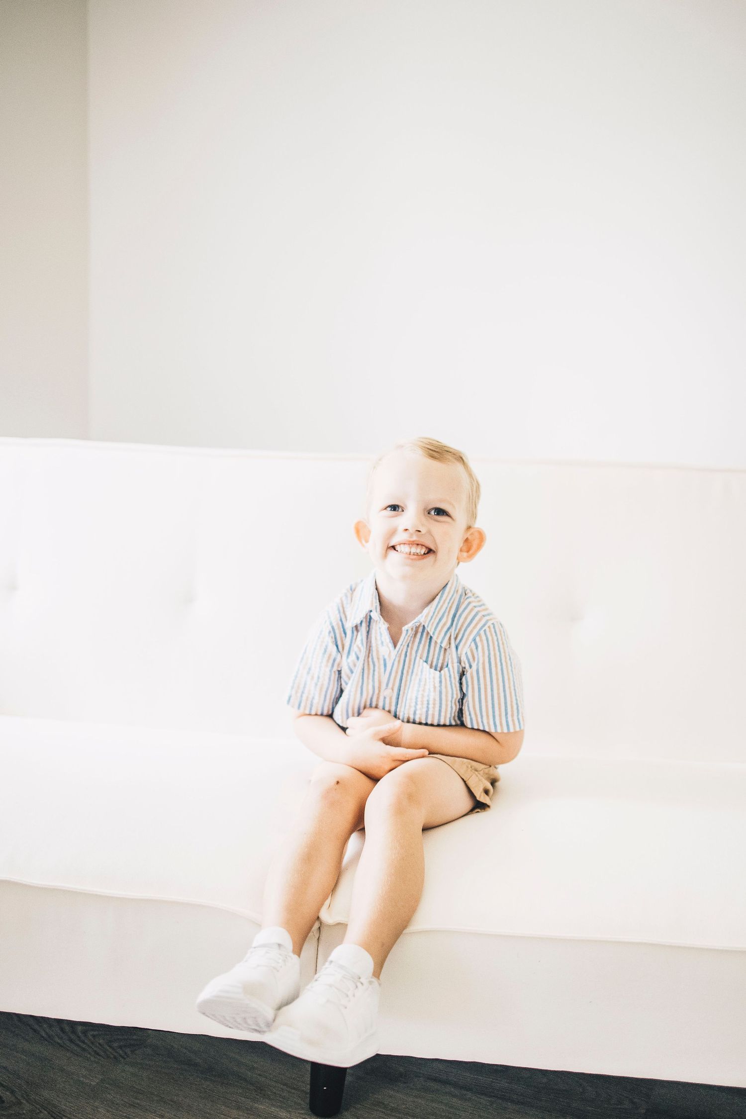 Cheerful young boy in striped shirt sitting on white couch smiling.