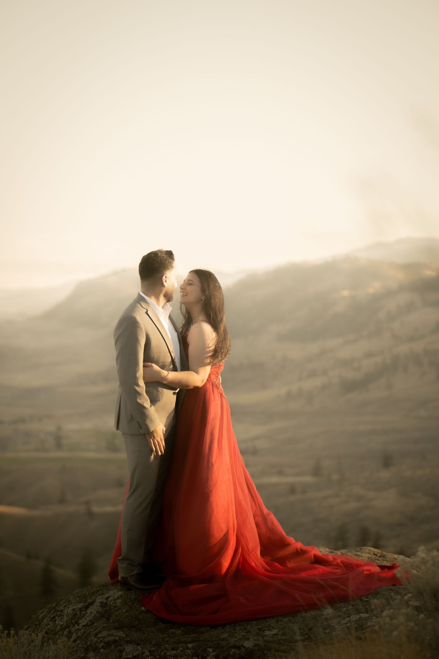 Romantic couple embraces on mountain overlook with flowing red dress against misty landscape at sunset.