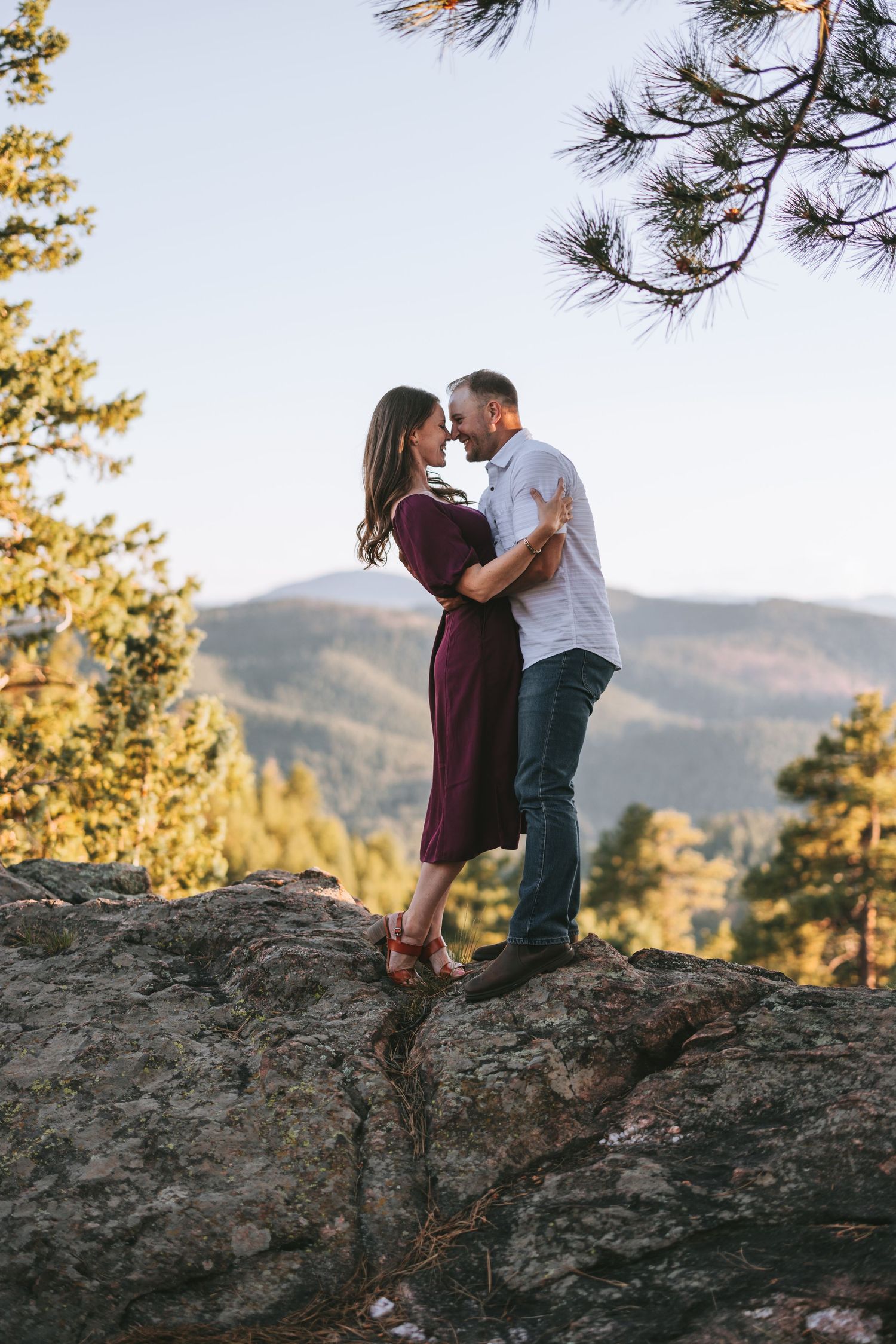 A couple embraces romantically on a mountain cliff during sunset with pine trees in the background.