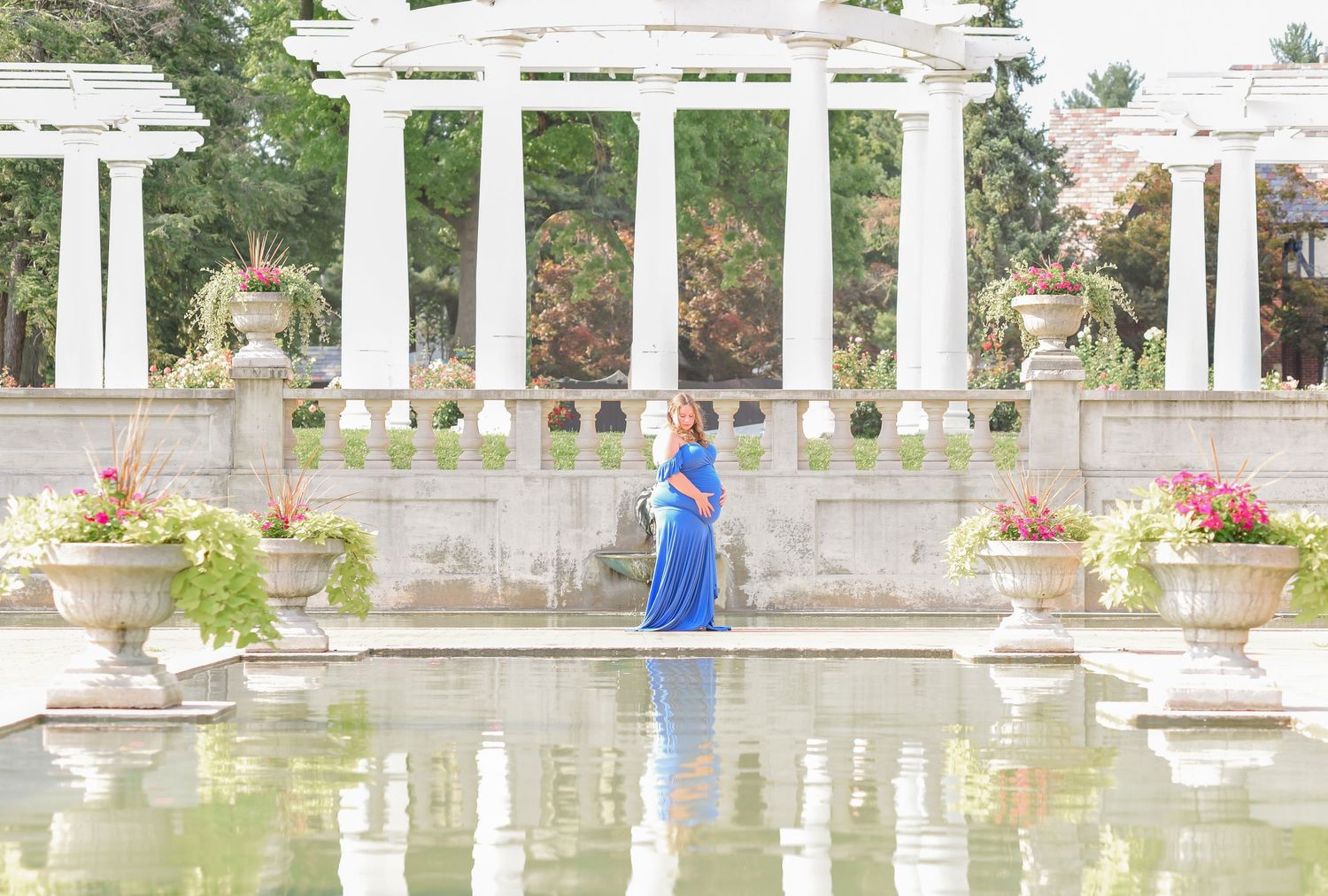 A person in a blue dress poses by an ornate reflecting pool with white columns and flower arrangements.