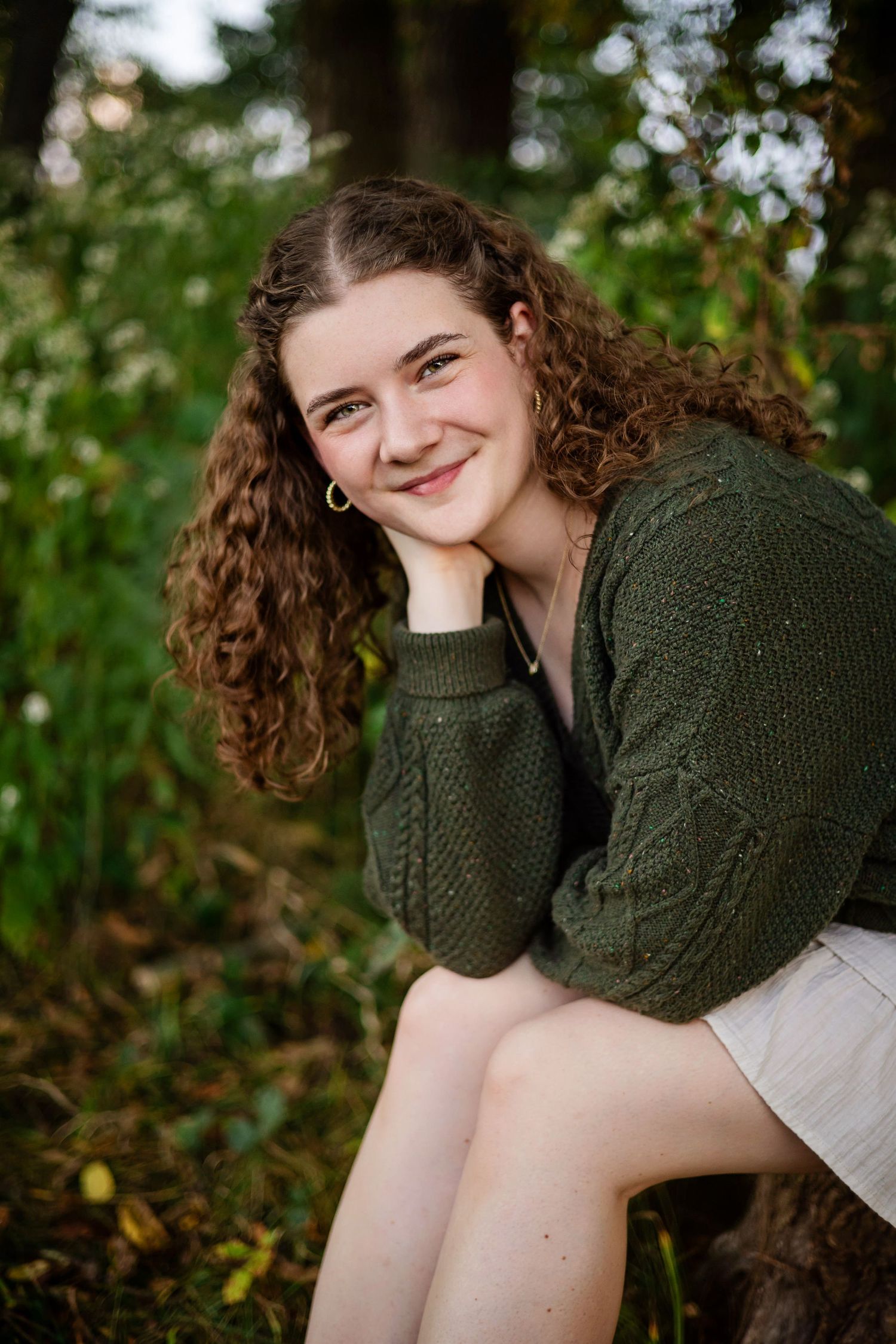 A portrait of someone with curly brown hair wearing a green sweater sitting outdoors with a natural background.