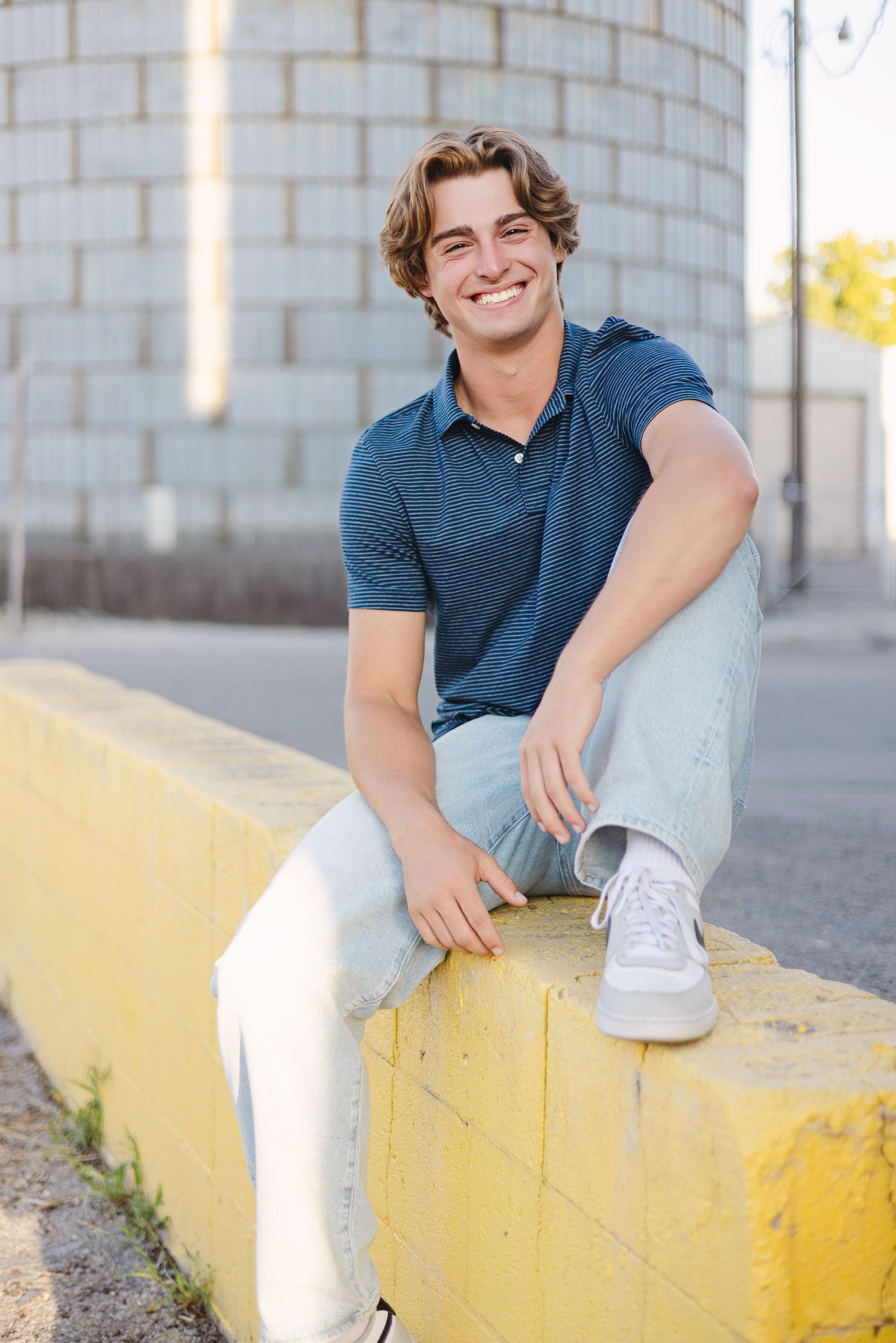 Casual portrait of someone sitting on yellow concrete barrier wearing navy polo shirt and light jeans with white sneakers.