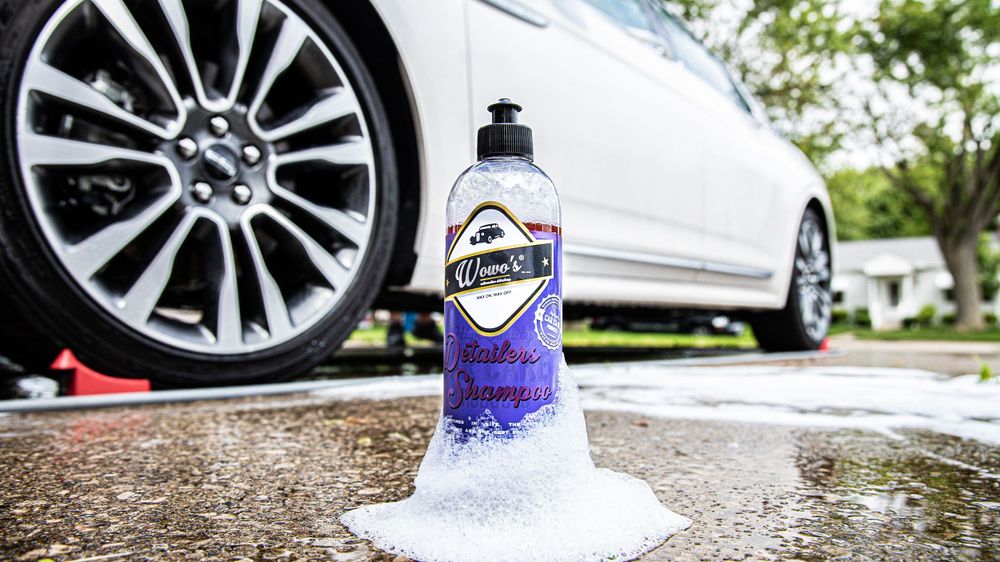 Purple car cleaning product bottle displayed with foam on ground near vehicle wheel.