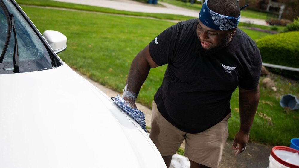Professional detailing service provider cleaning white car exterior on residential driveway.