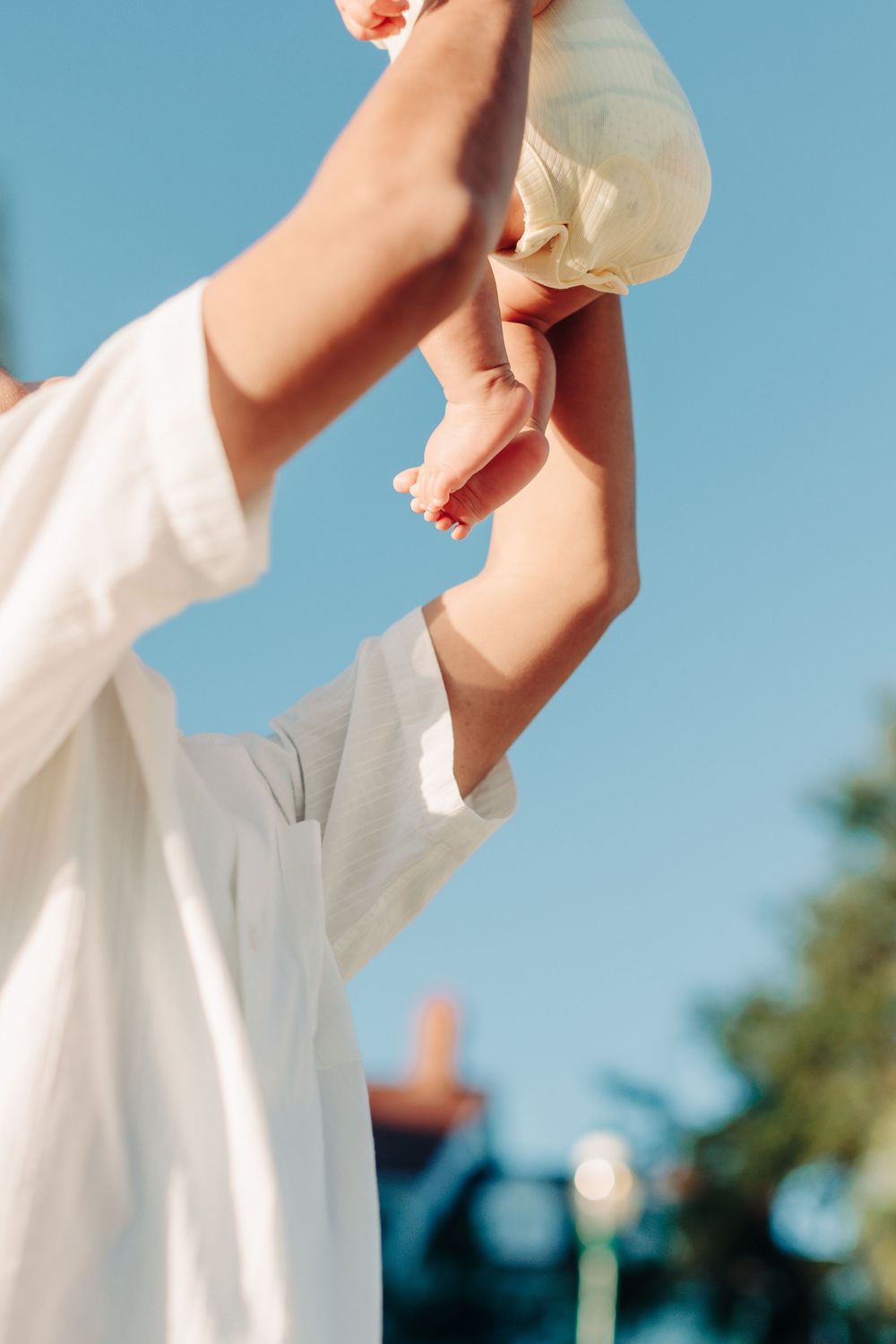 Hands lift a baby joyfully against a bright blue summer sky.