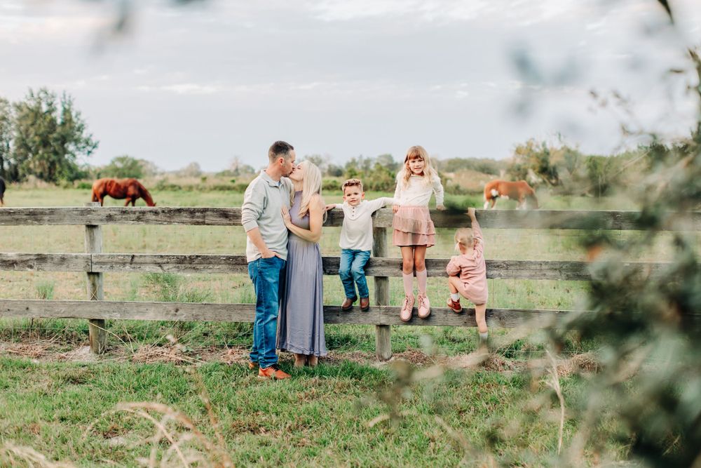 Intimate view through the trees of a family near horse ranch with mom and dad kissing and kids climbing on the fence with horse in the back