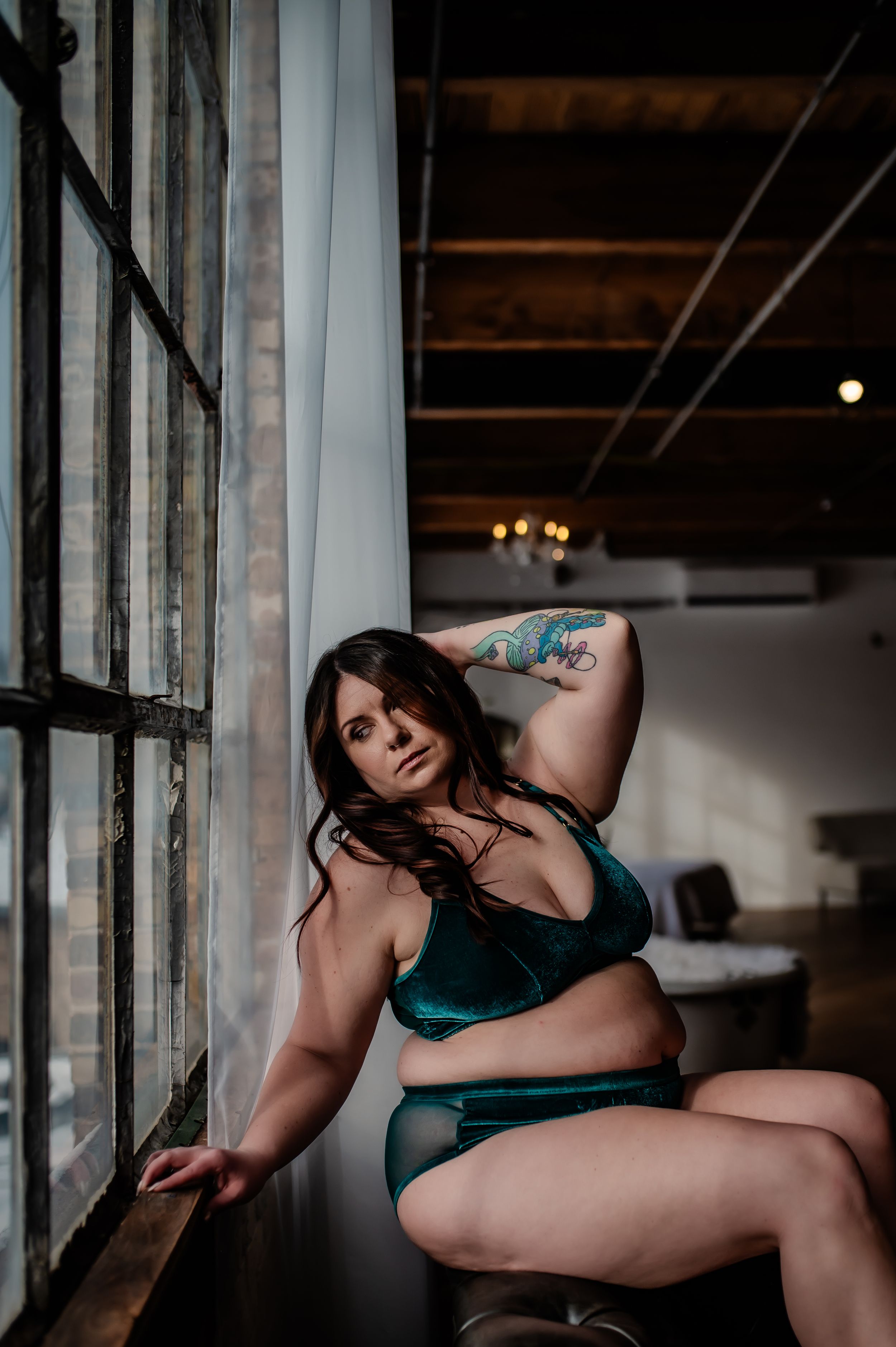 A portrait in moody lighting features someone in teal lingerie posing next to a window in an intimate boudoir setting.