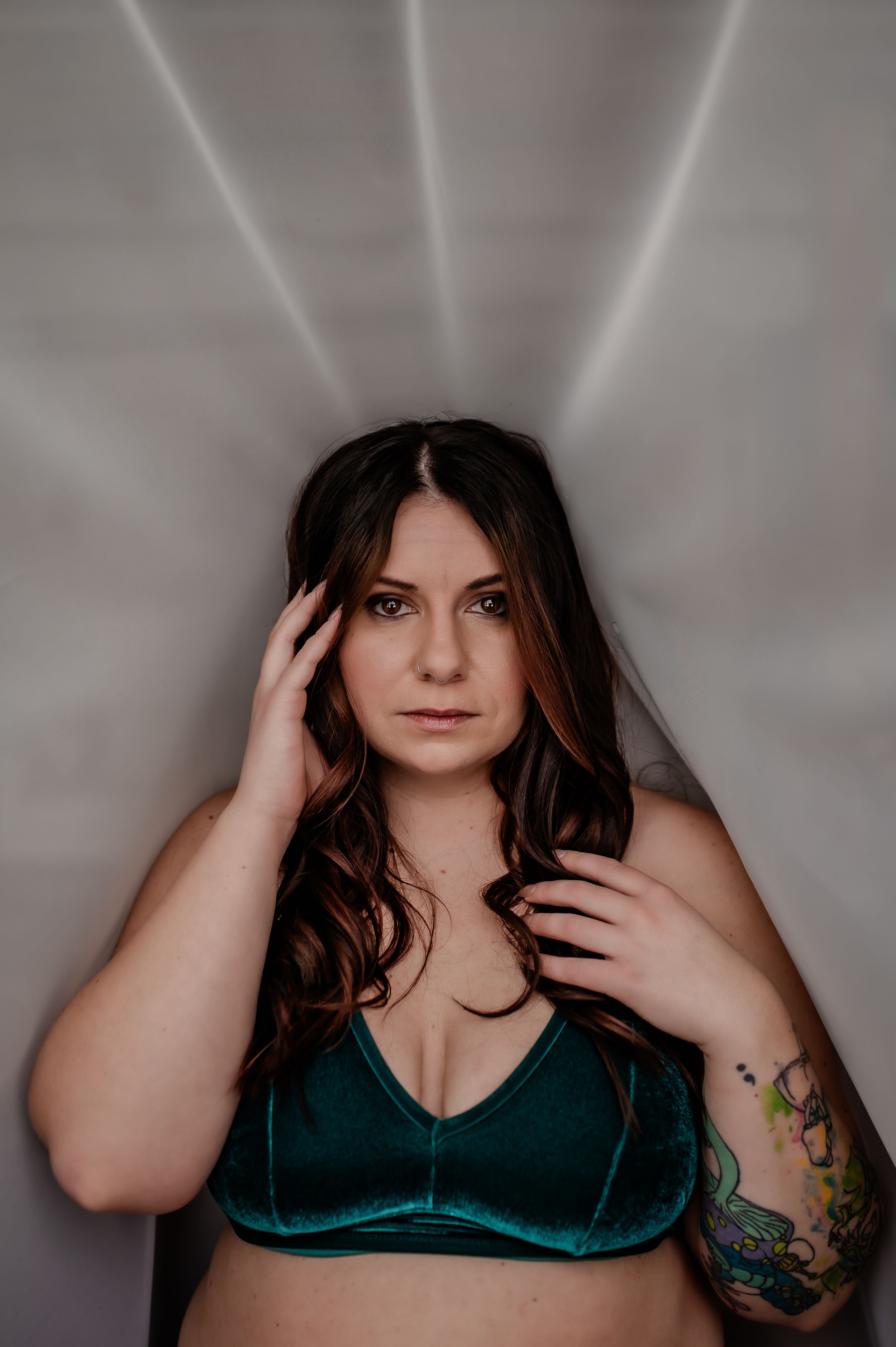 Portrait against gray background with dramatic lighting features person in emerald velvet bralette with tattoo on arm.