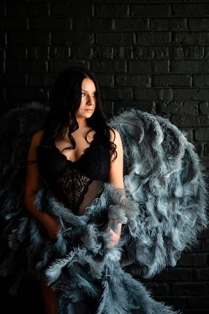 Dark-haired woman in black lace lingerie poses with large blue-gray feathered angel wings against a dark brick wall.