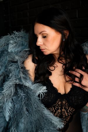 Woman in black lace lingerie draped with a blue feather coat, posing against a dark brick wall.