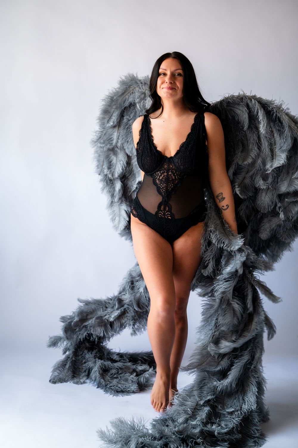 Woman in black lace bodysuit posed with large gray feather wings in a studio setting.