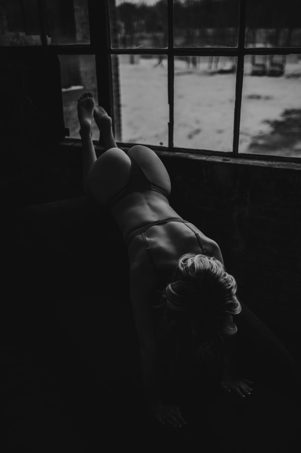 A black and white artistic boudoir photograph of a figure reclining by a window in moody lighting.