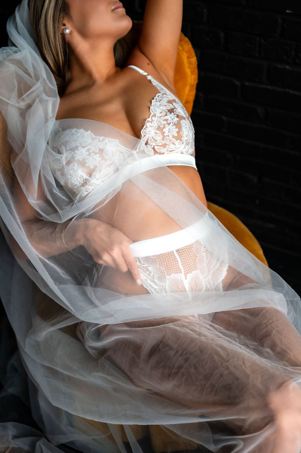 Woman in white lace lingerie and sheer robe reclining gracefully against a dark brick wall background.