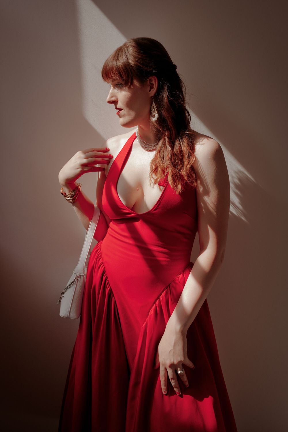 Woman in elegant red halter gown holding white clutch, posing in dramatic sunlight against neutral background.