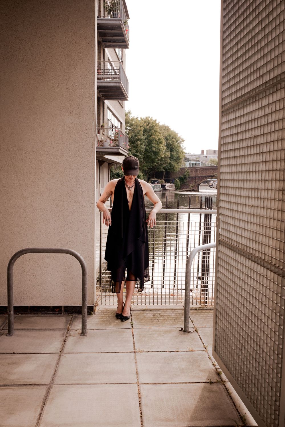 Woman in black dress and hat poses on urban balcony with metal railings and apartment buildings in background.
