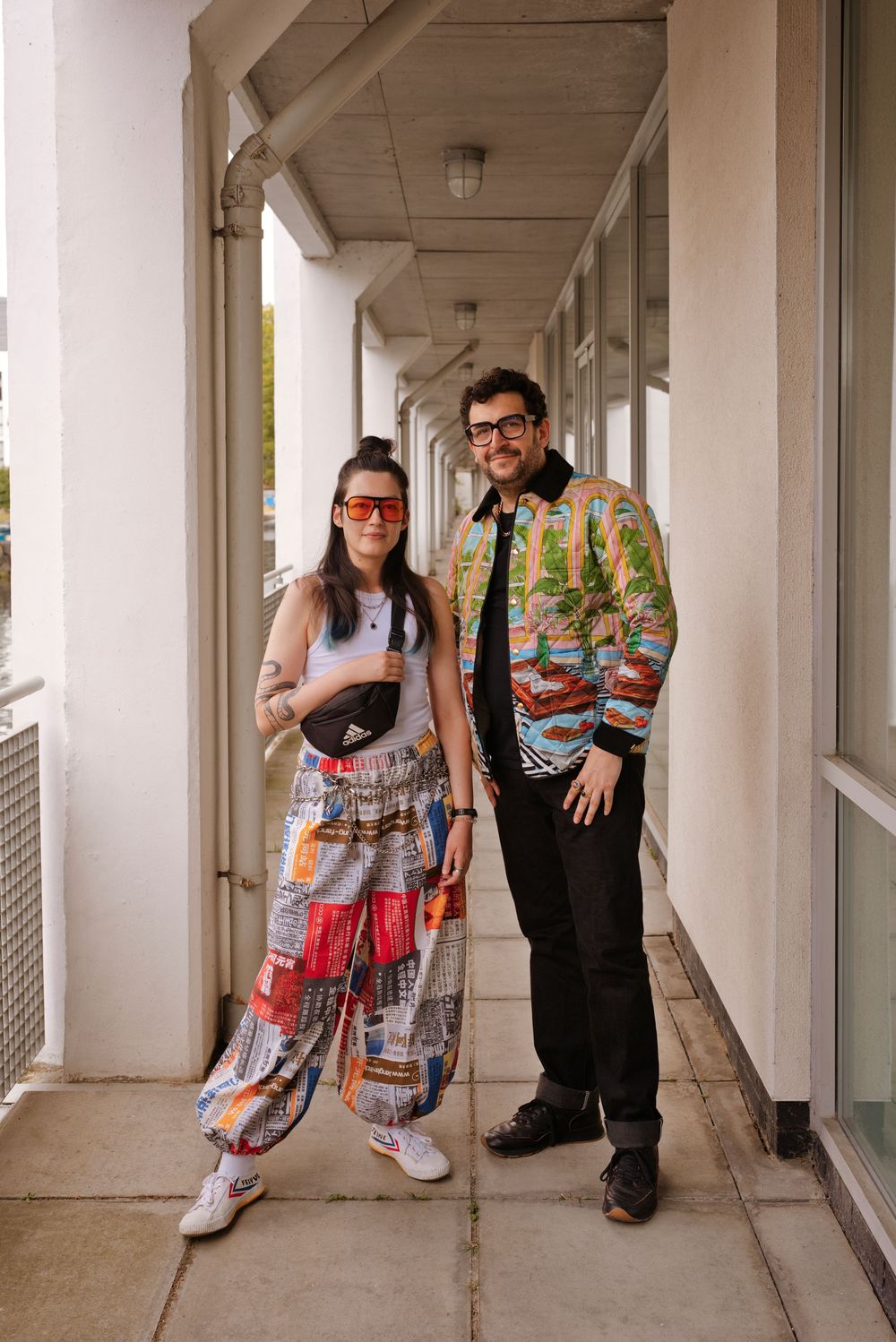 Two stylish people posing on a building corridor, wearing colorful patchwork pants and a vibrant printed bomber jacket.