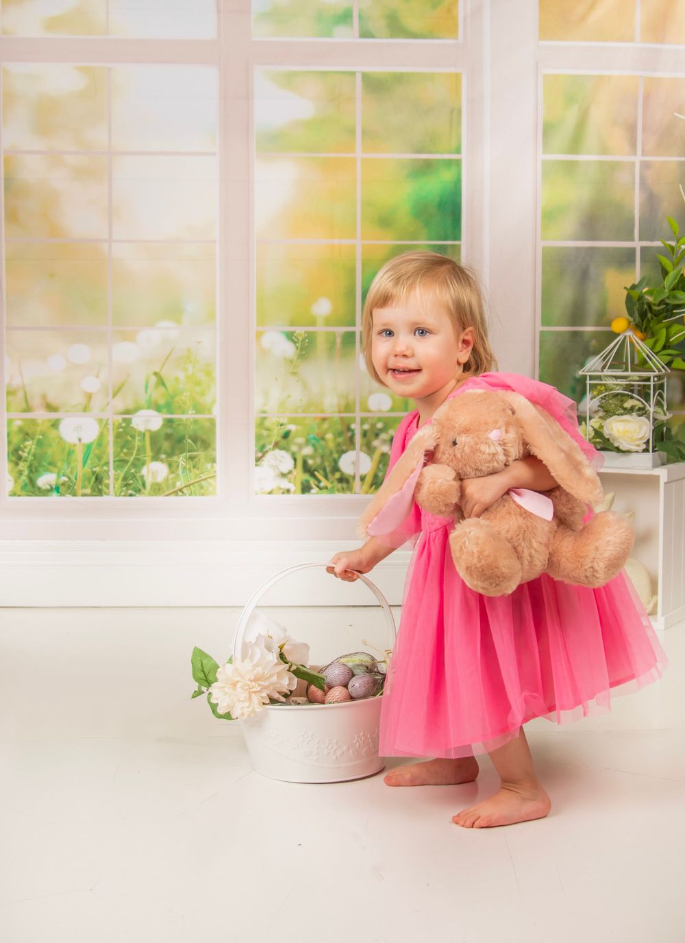 A child in a pink tutu dress holds a teddy bear while standing by a bright window.