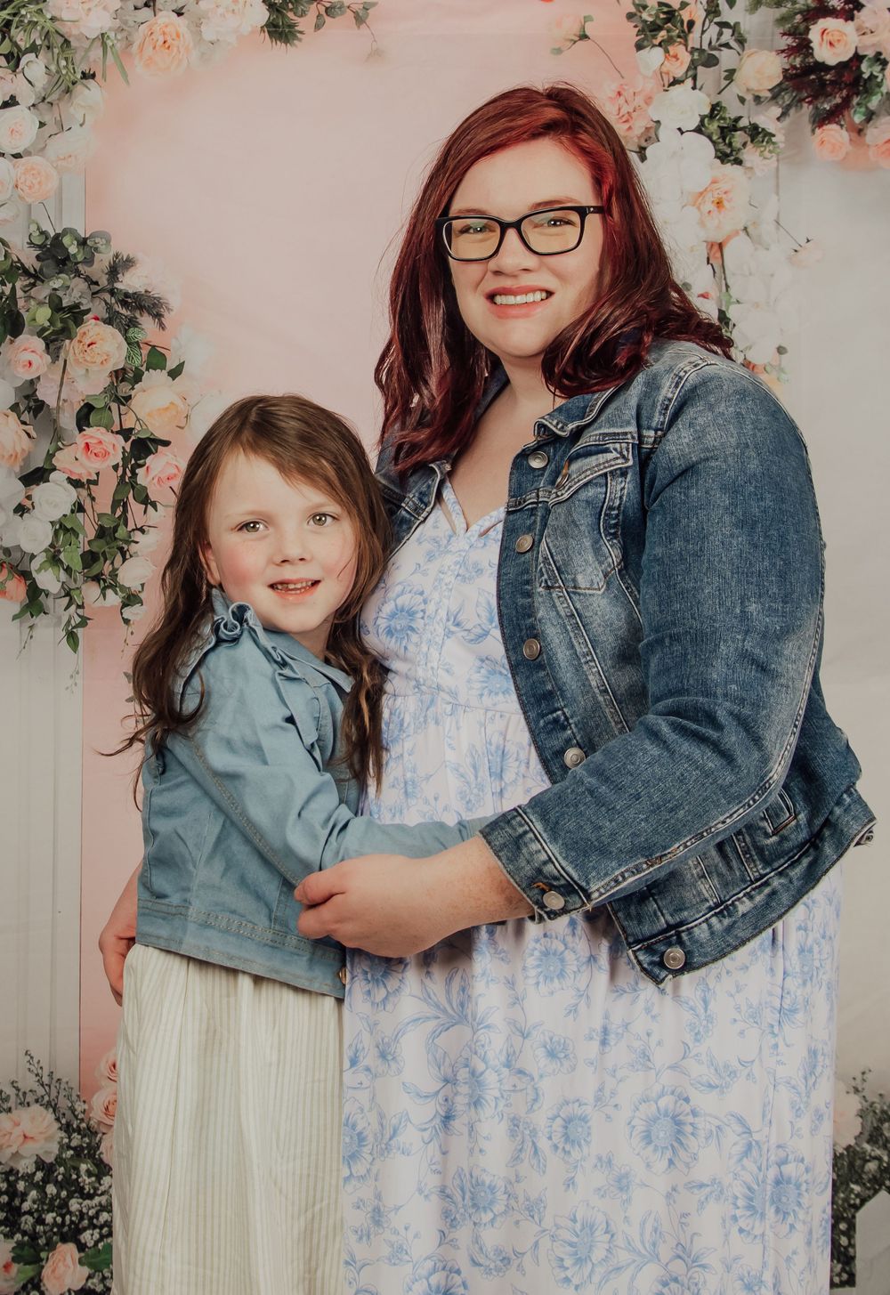 Two figures pose against a floral backdrop wearing denim jackets and spring outfits in a studio portrait setting.
