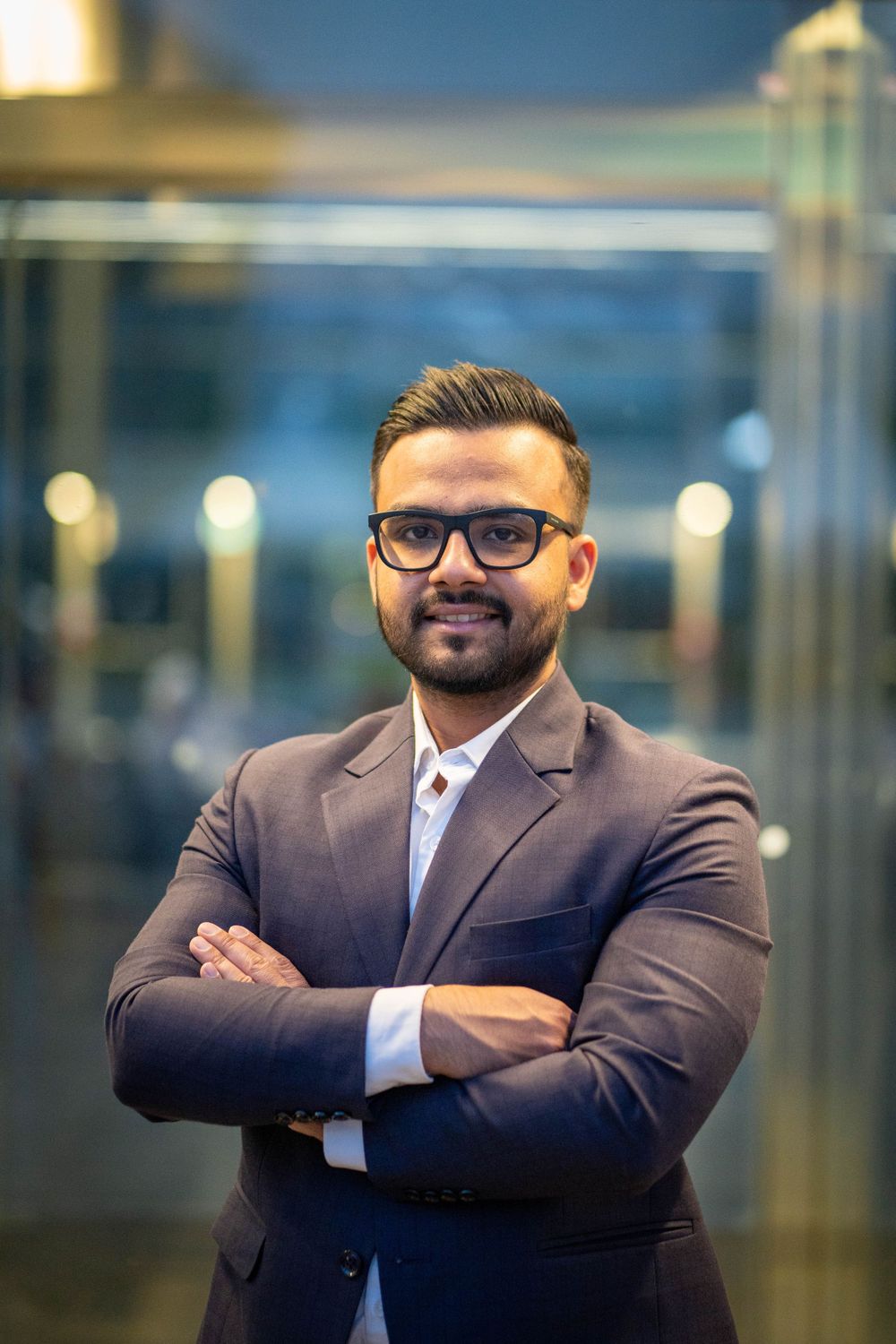 Professional in brown suit and glasses poses with crossed arms in corporate setting.