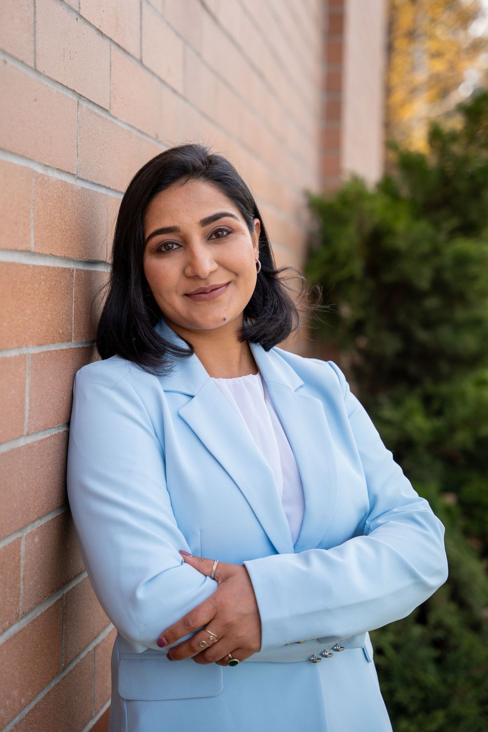 Professional in light blue blazer and white top poses against brick wall outdoors.