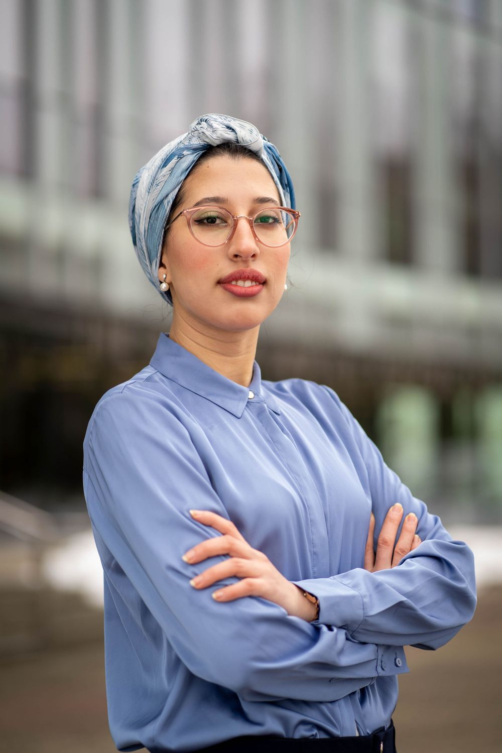 Professional wearing a light blue button-down shirt, blue headscarf, and round glasses poses with arms crossed.