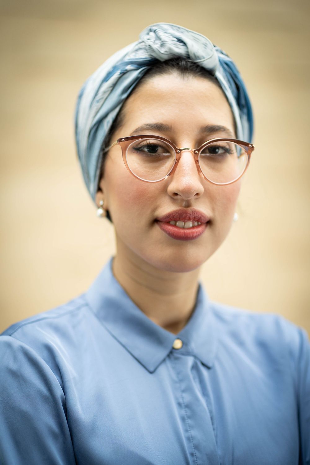 Close-up portrait of someone in a blue collared shirt, light blue headscarf and wire-frame glasses.