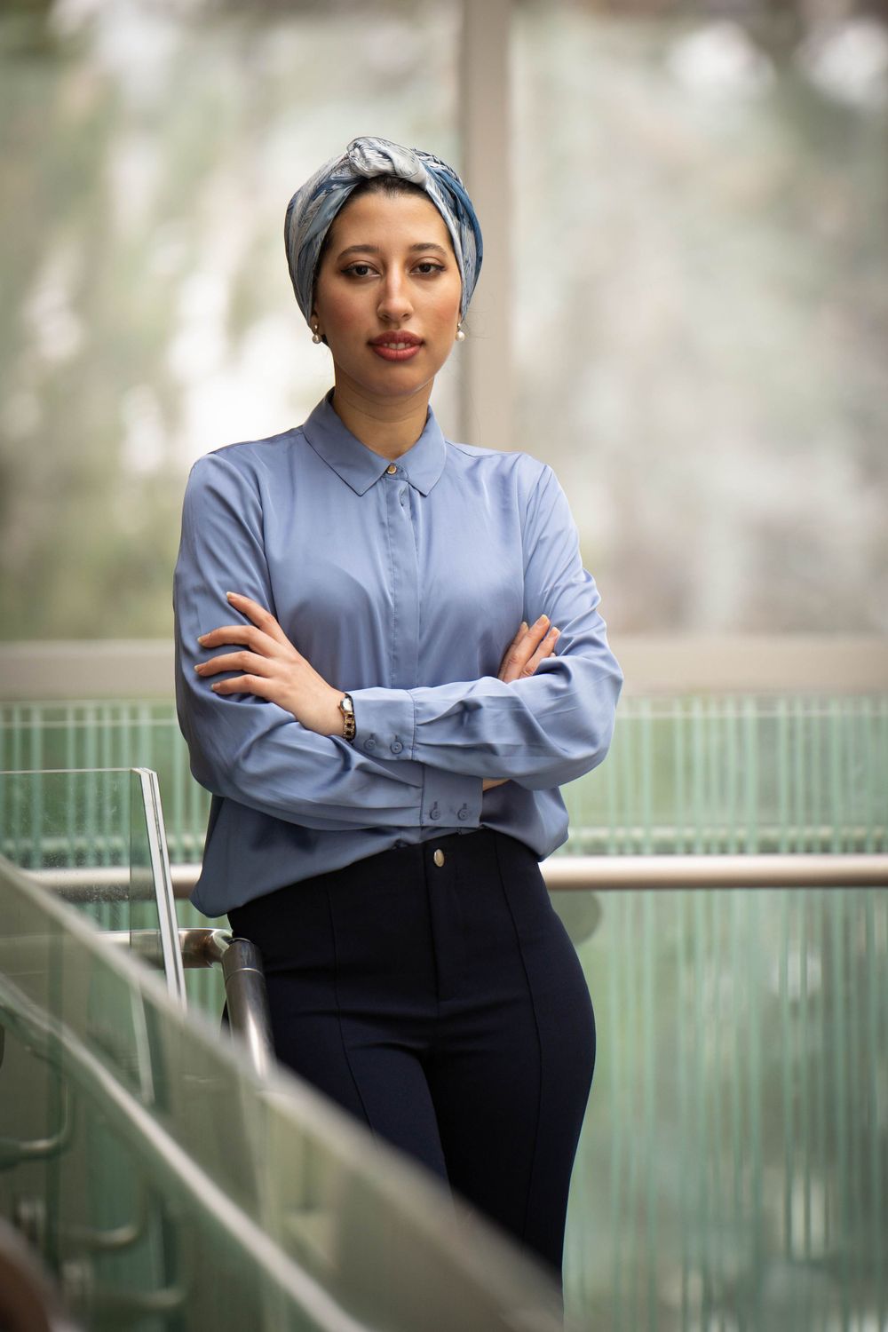 Professional in blue shirt and navy pants poses confidently by glass railing in modern building.