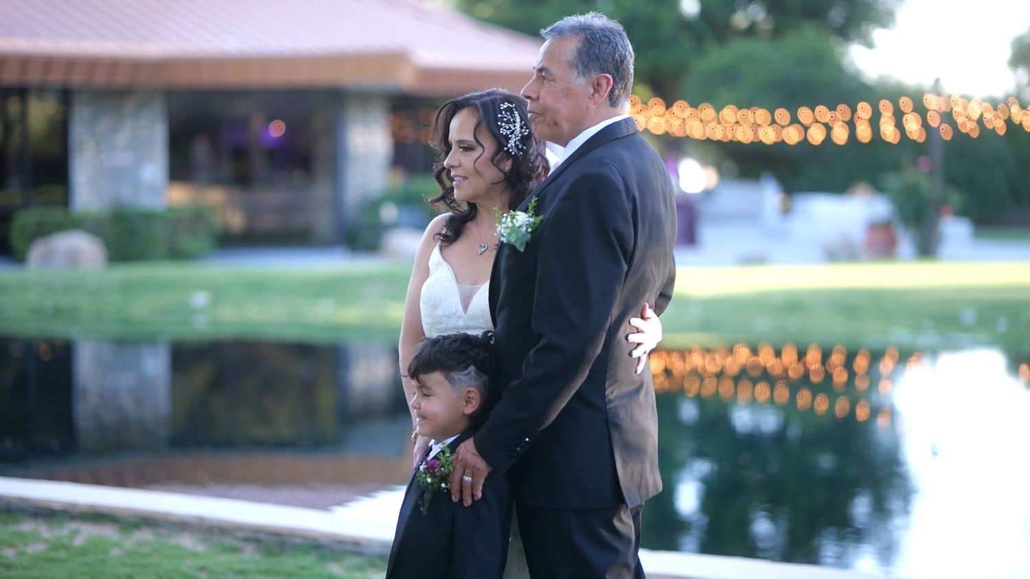 Wedding couple stands with young boy by a pond at dusk with twinkling lights in background.