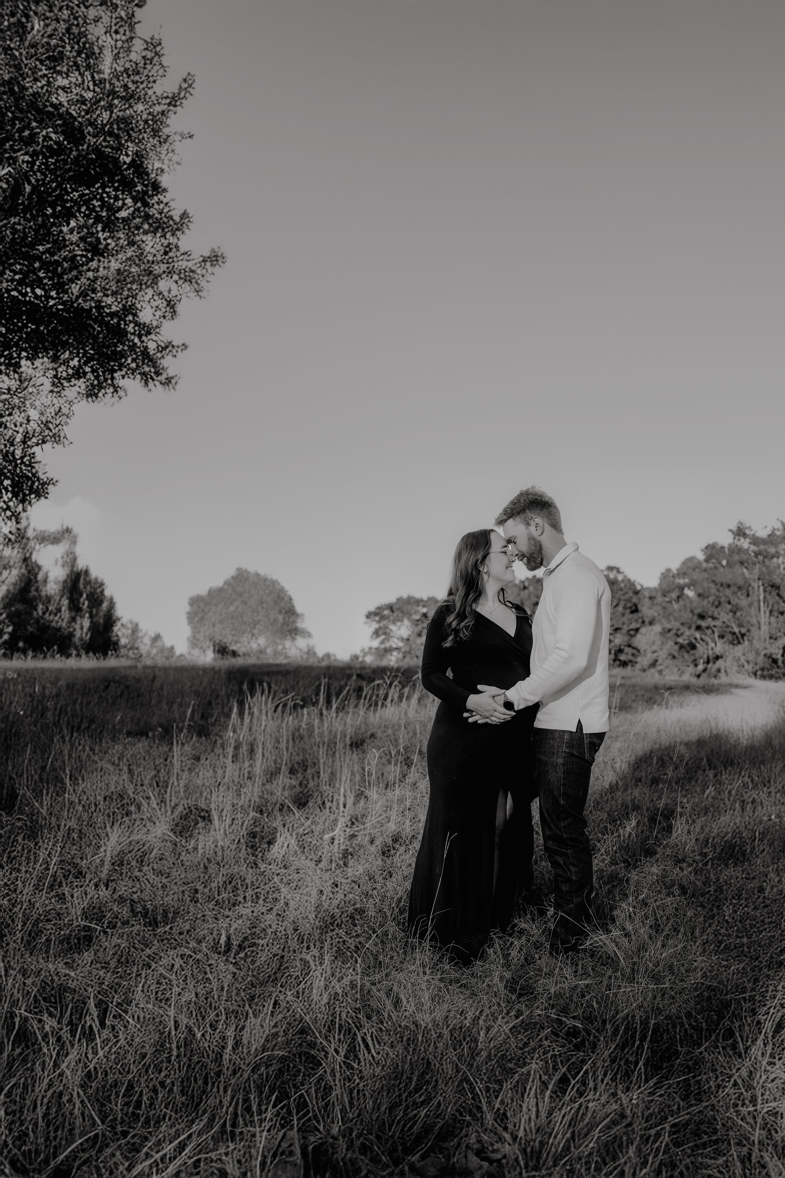 A couple embraces in a serene meadow with tall grass and trees silhouetted against an overcast sky in black and white.