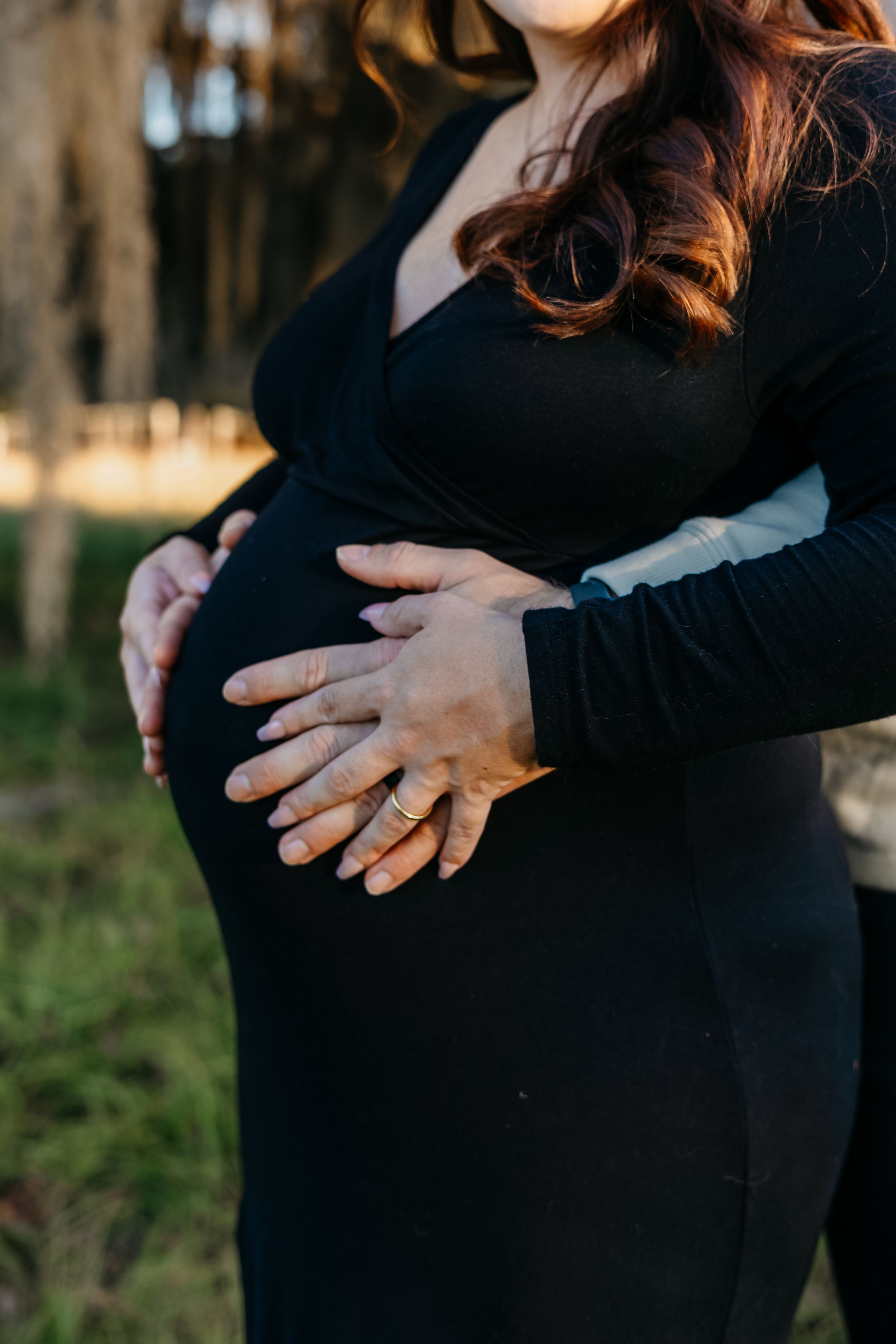 A pregnant woman in a black dress holds her baby bump while standing in a wooded outdoor setting.