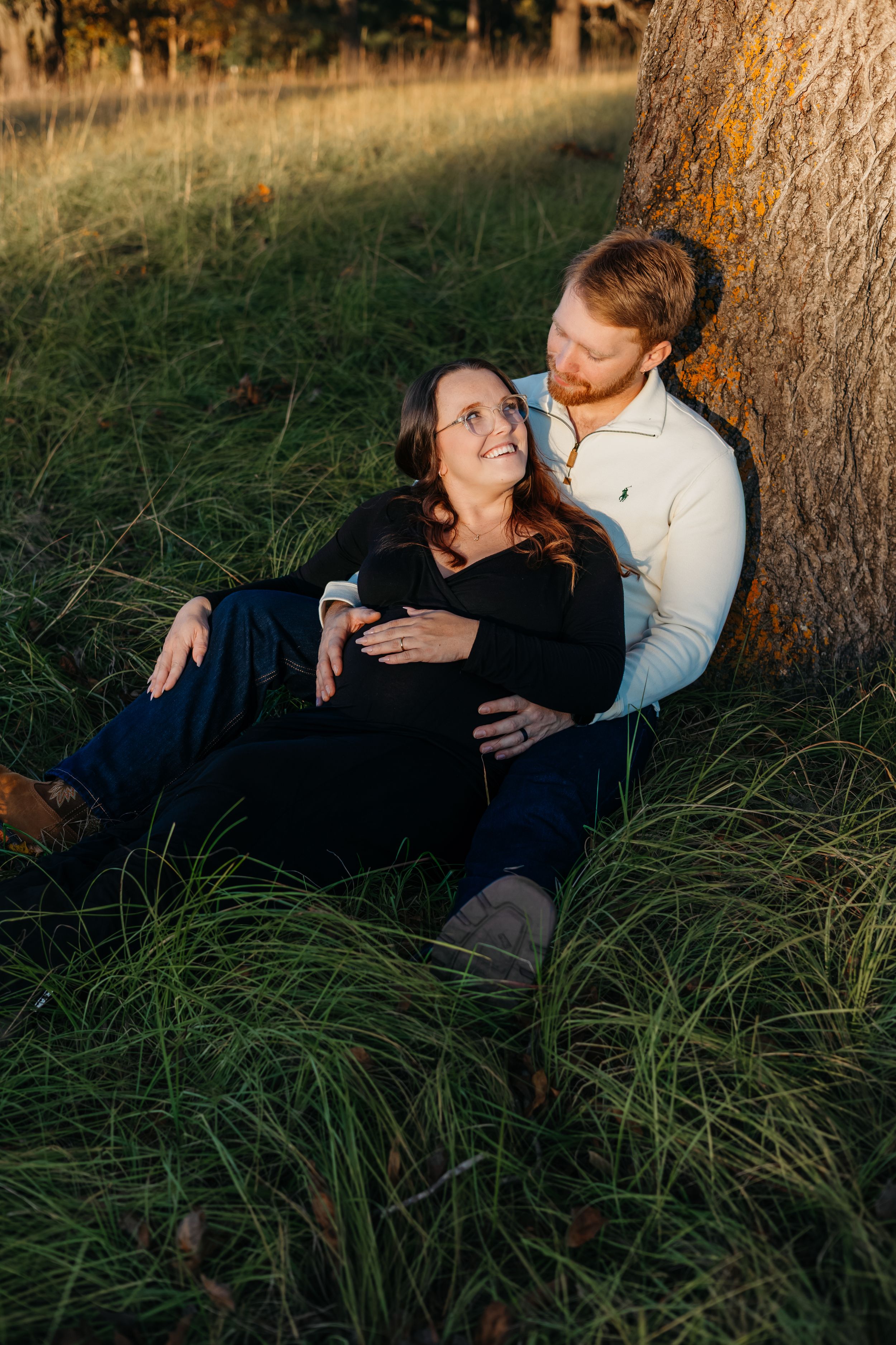 A couple relaxes together against a tree trunk in tall grass during golden hour sunset lighting.