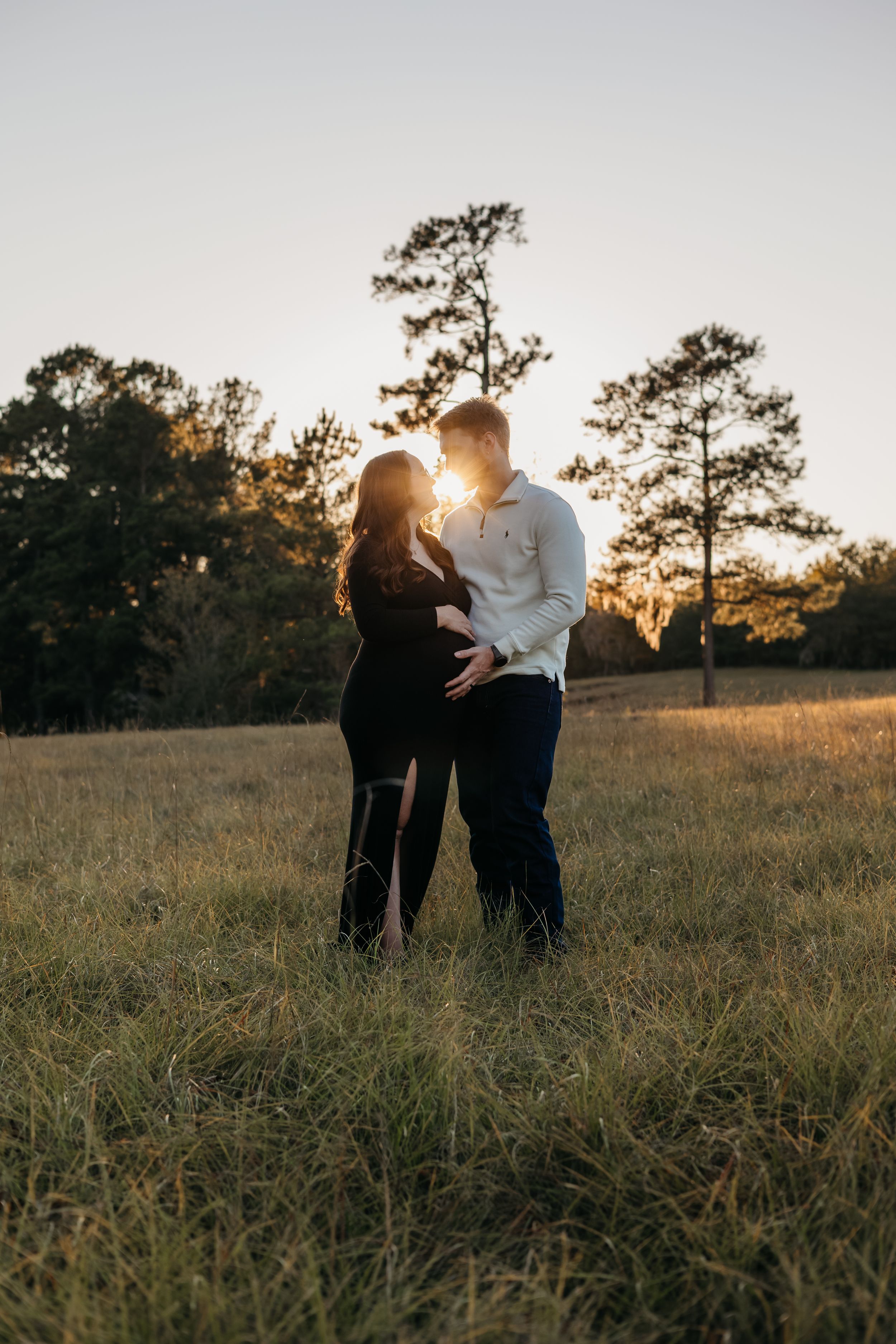 Silhouetted couple embraces in golden sunlit field at sunset with tall pine trees in background.